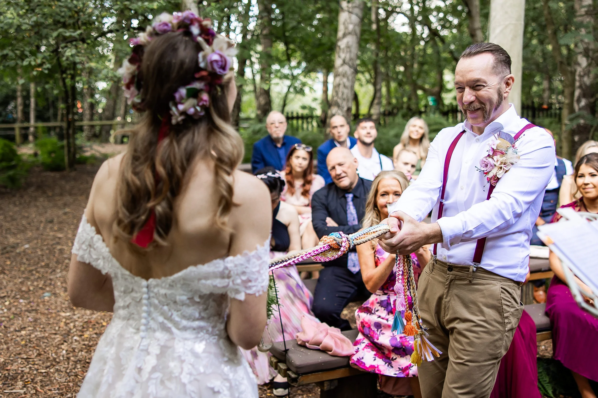 A wedding ceremony in an outdoor forest setting with a bride wearing a flower crown and a white lace dress, and a groom in a white shirt with suspenders and a floral boutonniere, holding a rope as part of a handfasting ritual.