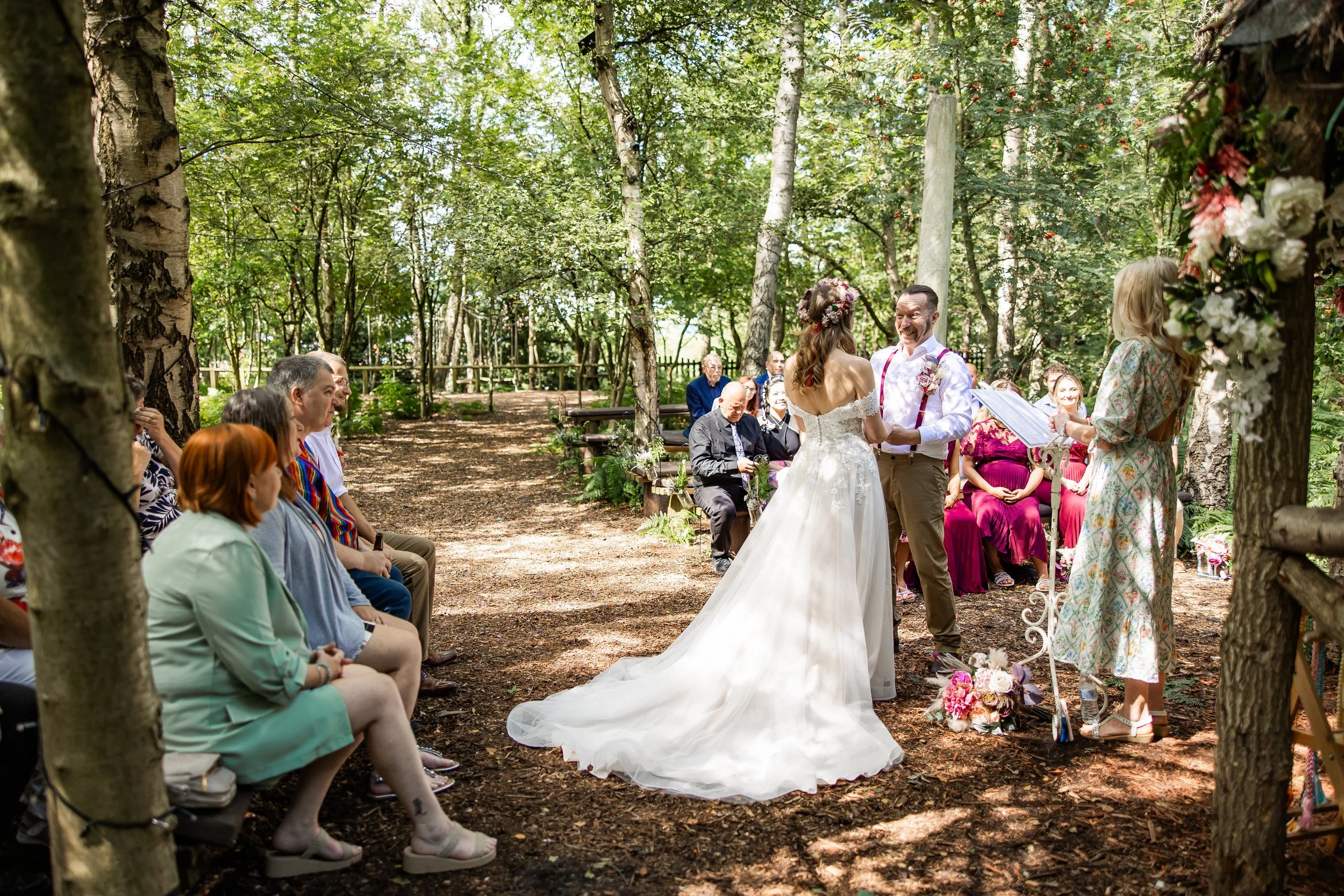 A wedding ceremony taking place outdoors in a wooded area with family and friends seated, the bride and groom holding hands and smiling in front of an officiant.