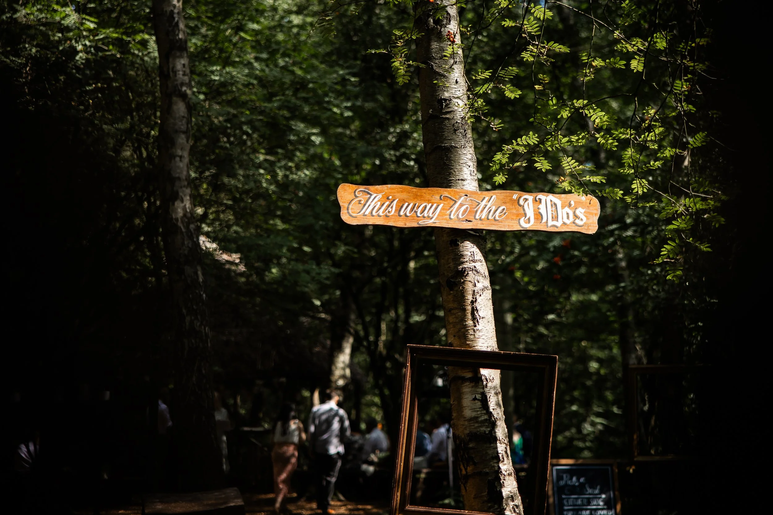 Wooden sign attached to a tree in a forest with the text 'This way to the I Do's' and people walking in the background.