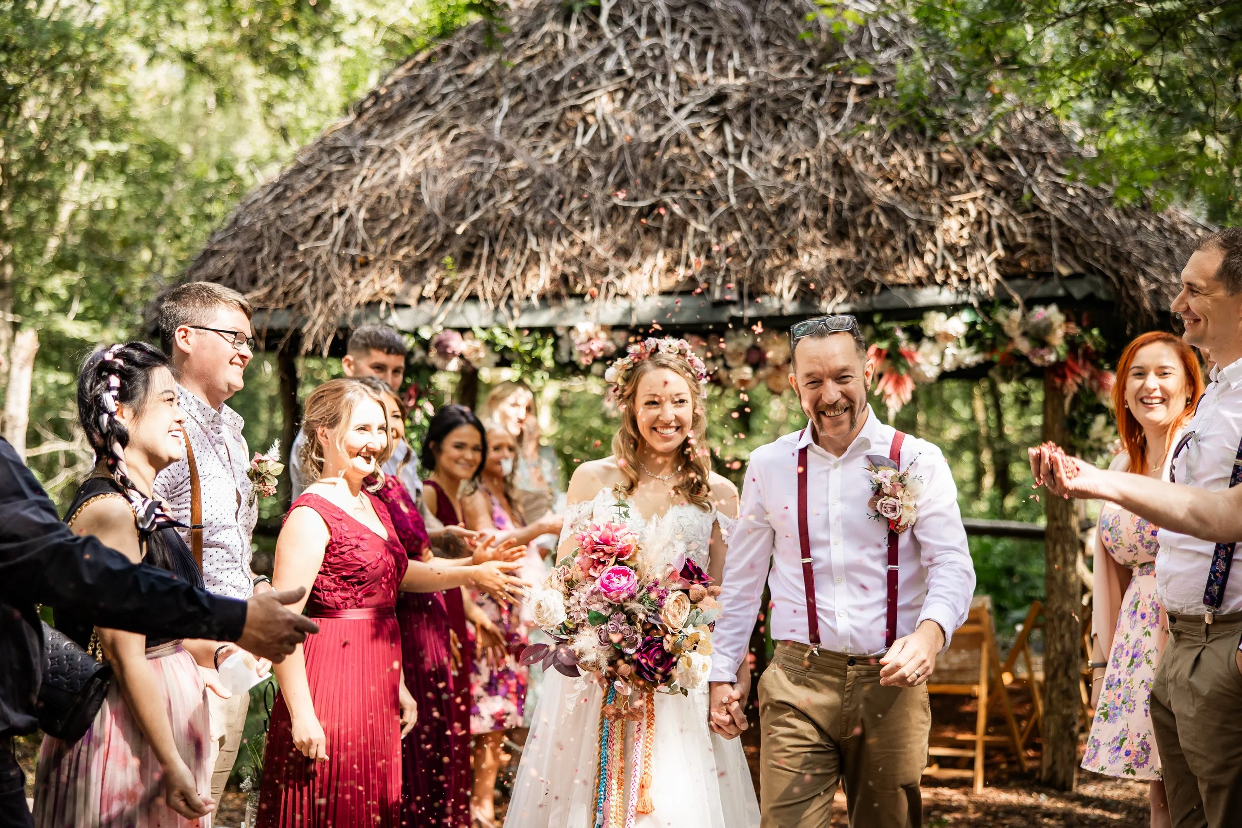 A wedding celebration outdoors with a bride and groom holding hands, surrounded by friends and family throwing flower petals, in front of a wooden shelter with a thatched roof, greenery, and floral decorations.