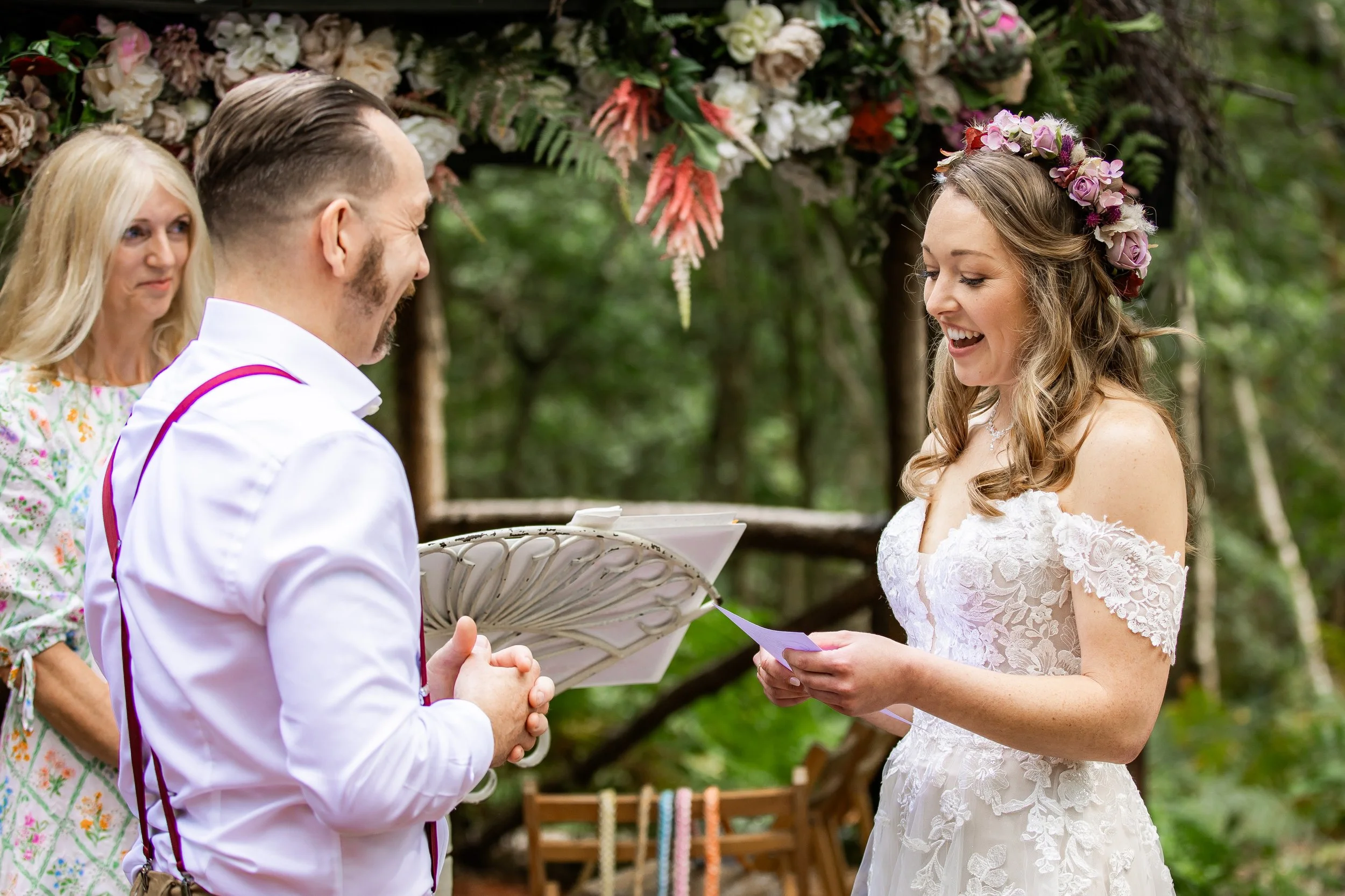 A bride and groom exchange vows during an outdoor wedding ceremony in a wooded area, with a female officiant standing behind them, and decorated with pink and purple flowers.