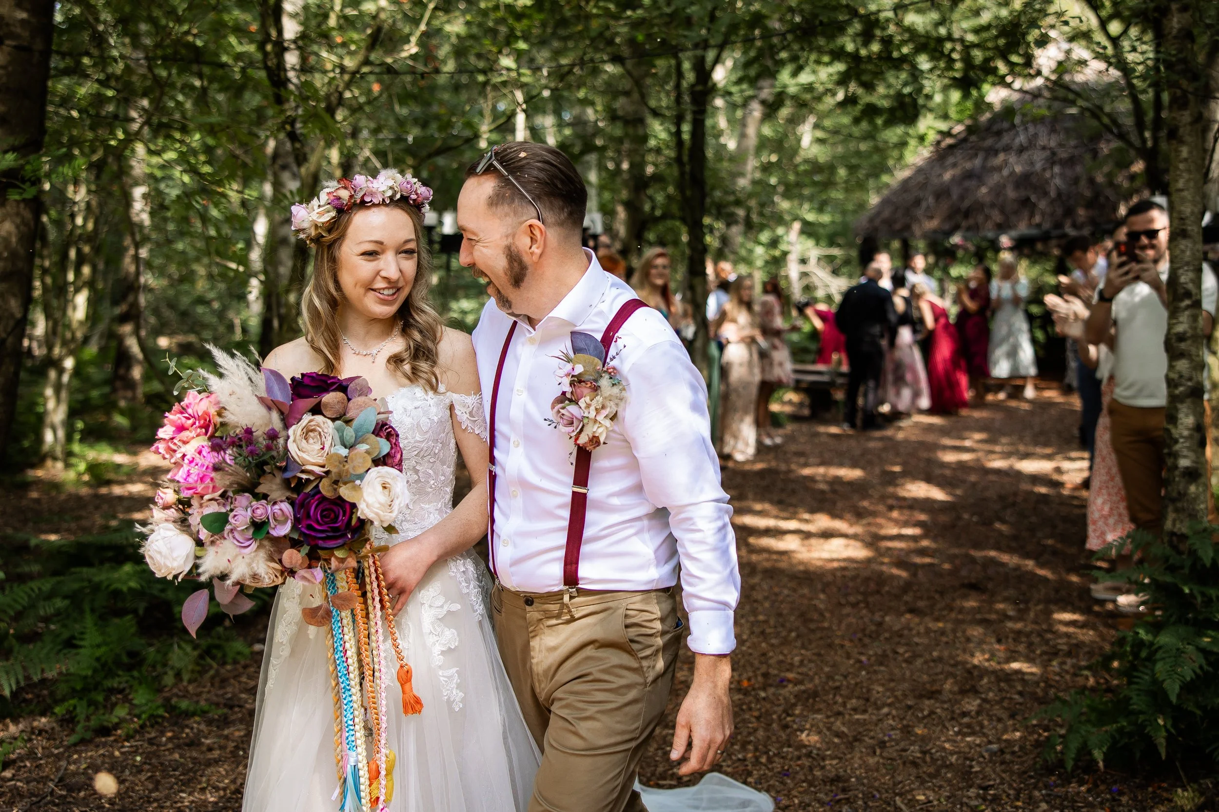 A bride and groom smile and laugh during their outdoor wedding in a wooded area, surrounded by guests.