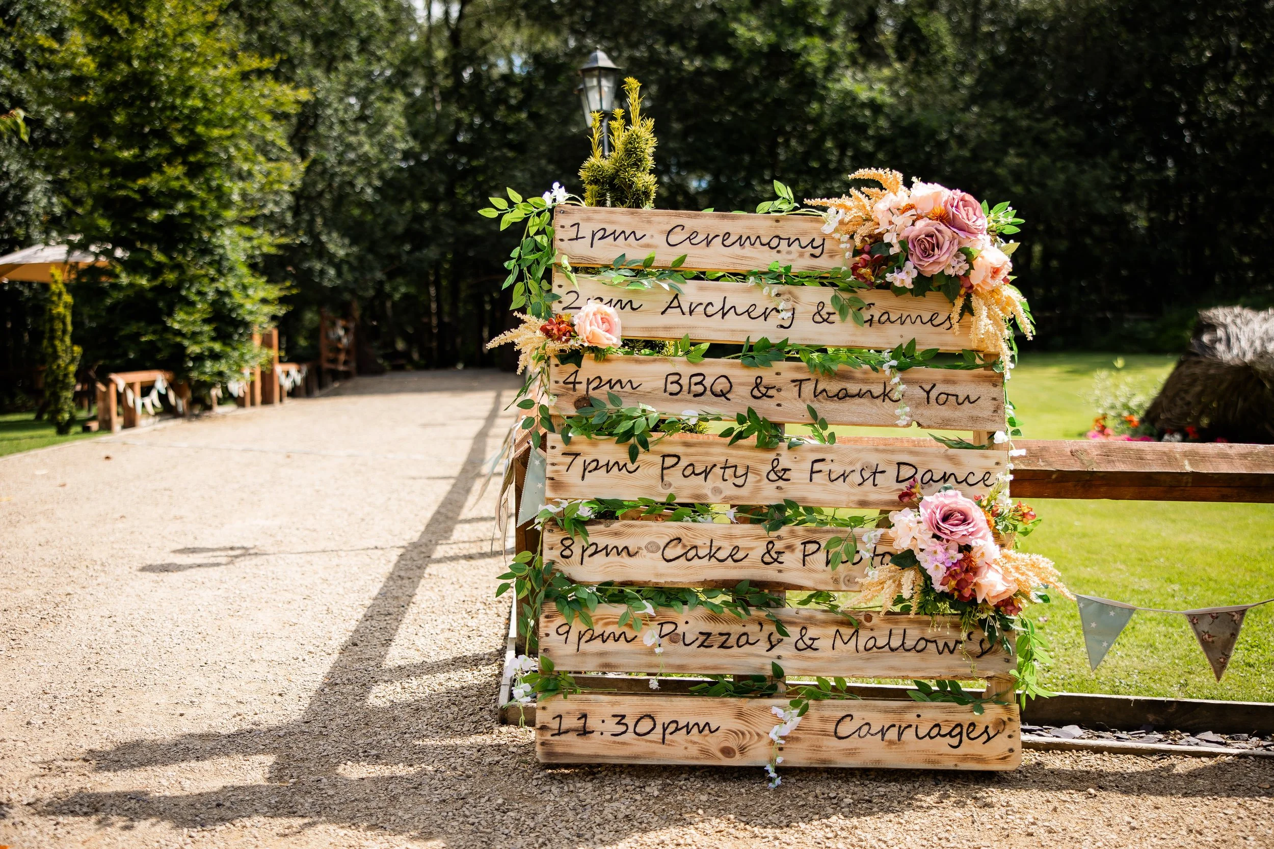 A wooden wedding schedule board decorated with flowers and greenery, listing events from 1 pm to 11:30 pm at an outdoor venue surrounded by trees and grass.