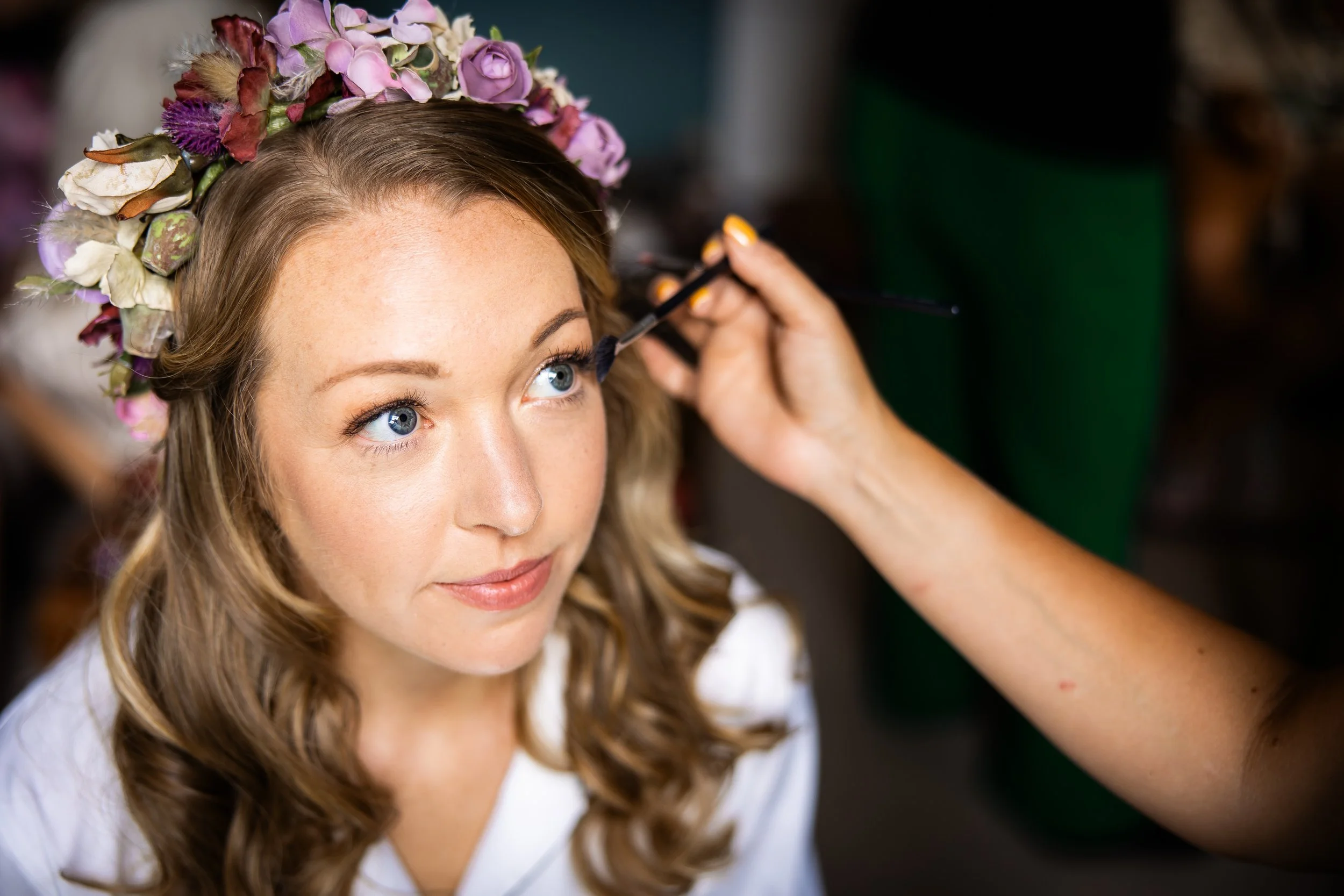 A woman with a floral crown of purple and white flowers getting her makeup done, with a makeup artist applying eye makeup.