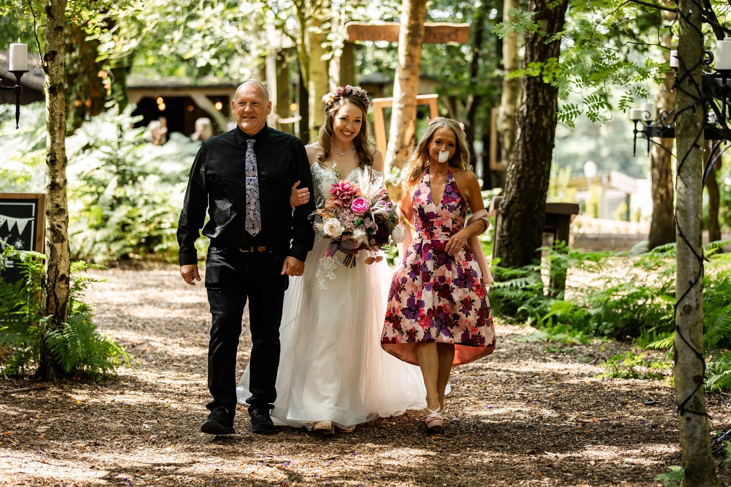 A bride in a white wedding dress holding a bouquet walks with two women and a man through a wooded outdoor area decorated for a wedding at Cheshire Woodland Weddings.