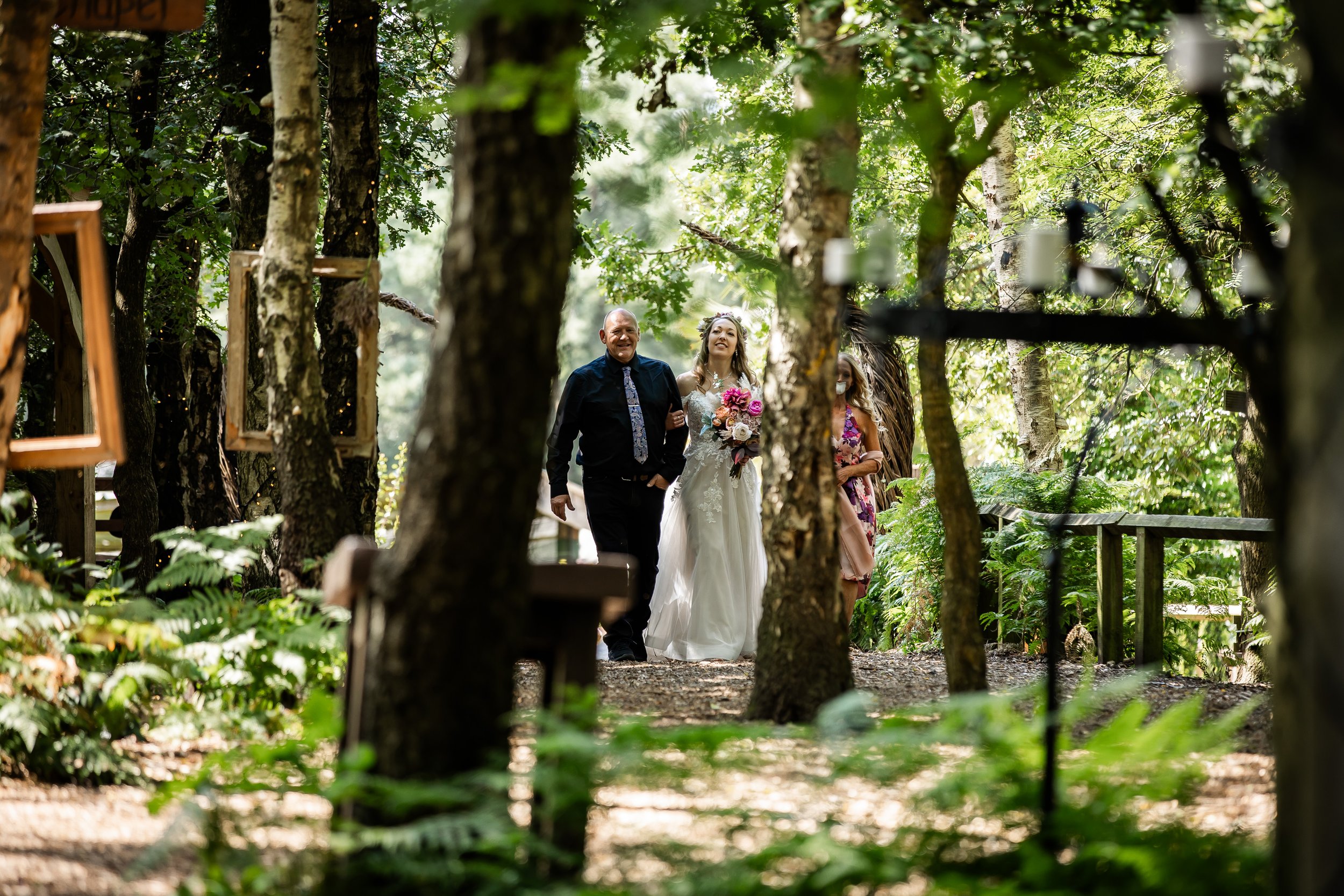 A bride in a white wedding dress holding a pink and white bouquet, walking down a wooded trail with a man at Cheshire Woodland Weddings.
