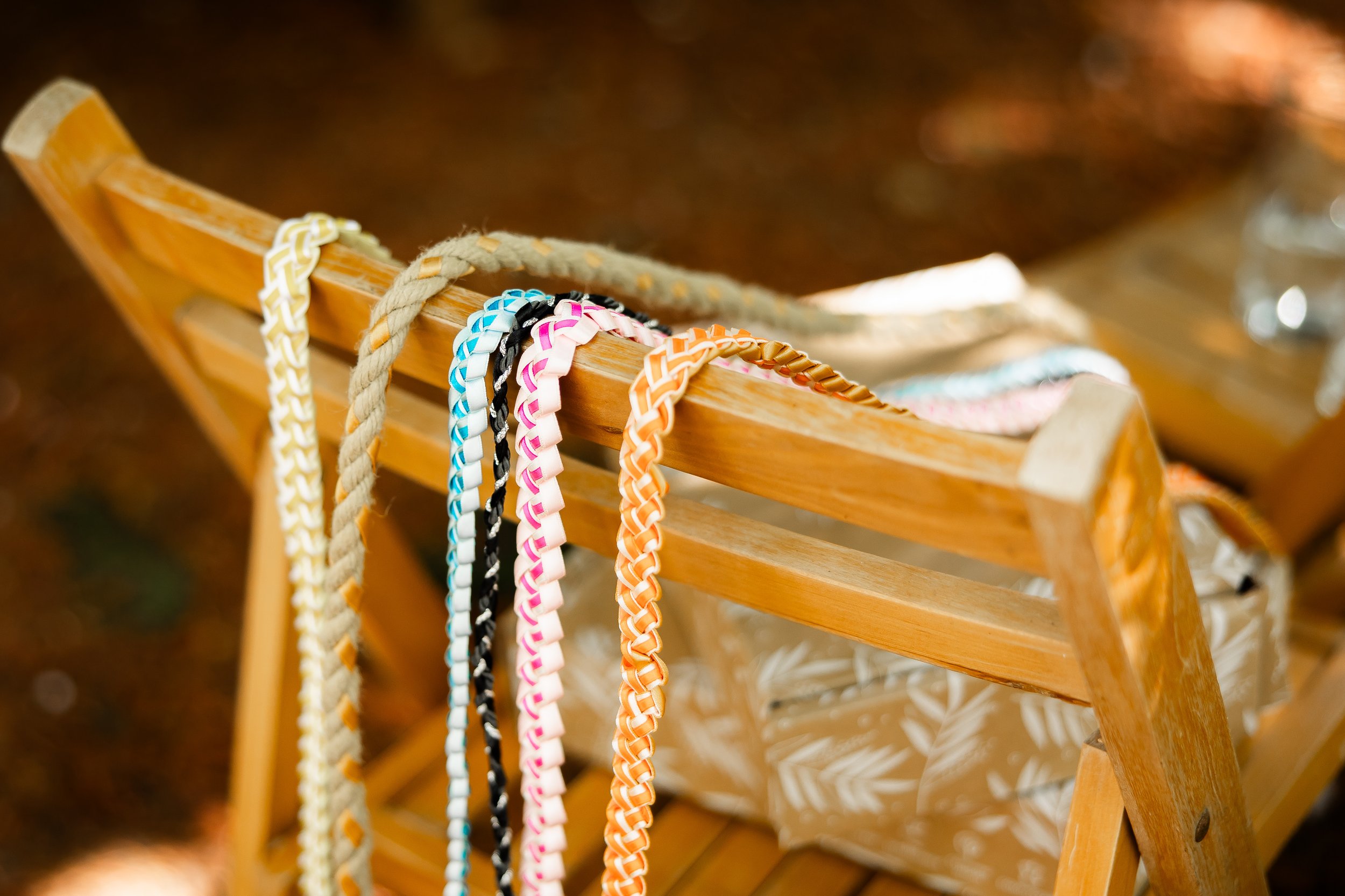 Colorful woven friendship bracelets hanging on a wooden chair with a patterned bag and a glass of water nearby in an outdoor setting.