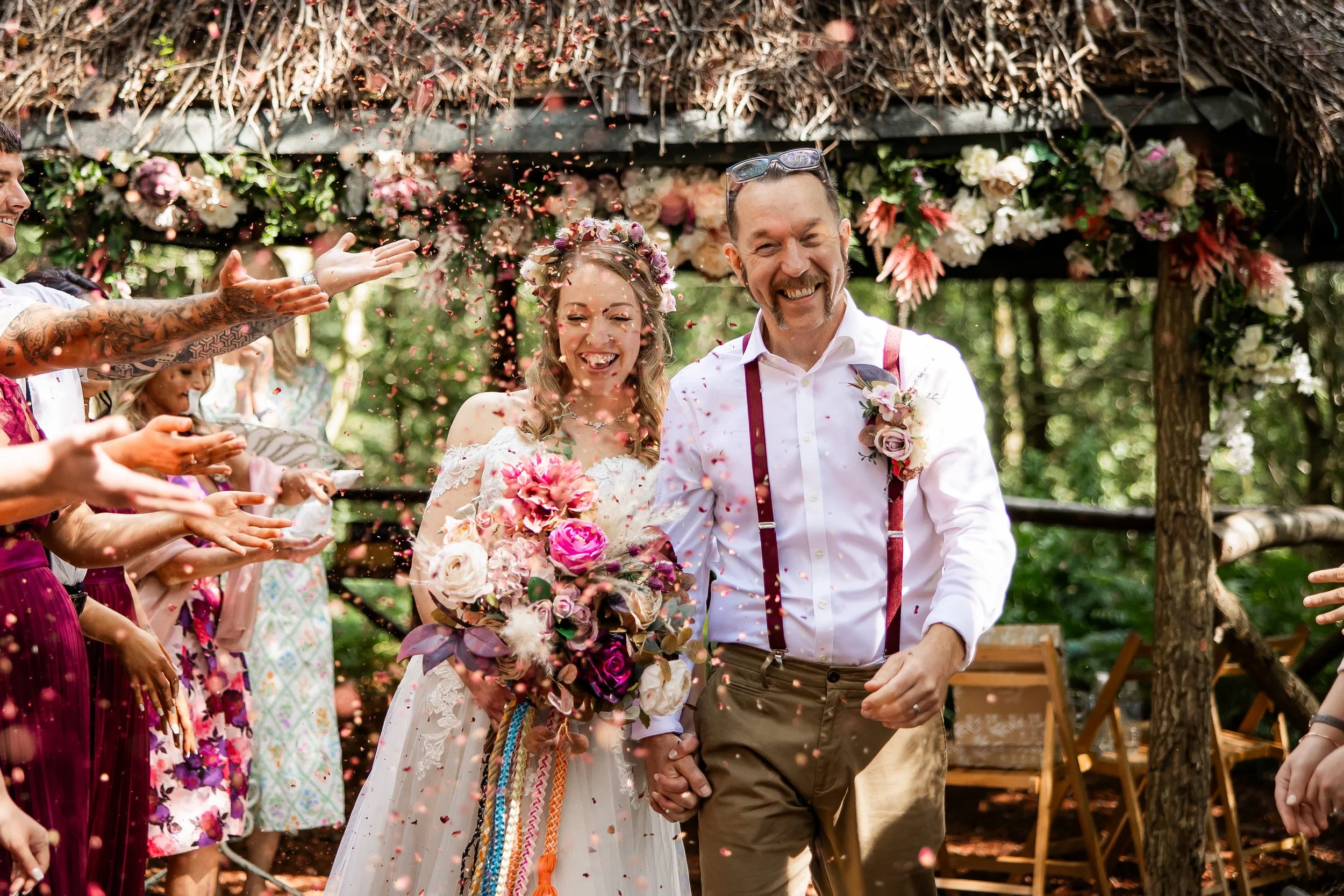 A newly married couple walks hand-in-hand through a shower of pink and peach confetti, smiling joyfully. The bride is in a white lace dress, holding a large bouquet of pink, purple, and white flowers. The groom is dressed in a white shirt with suspen