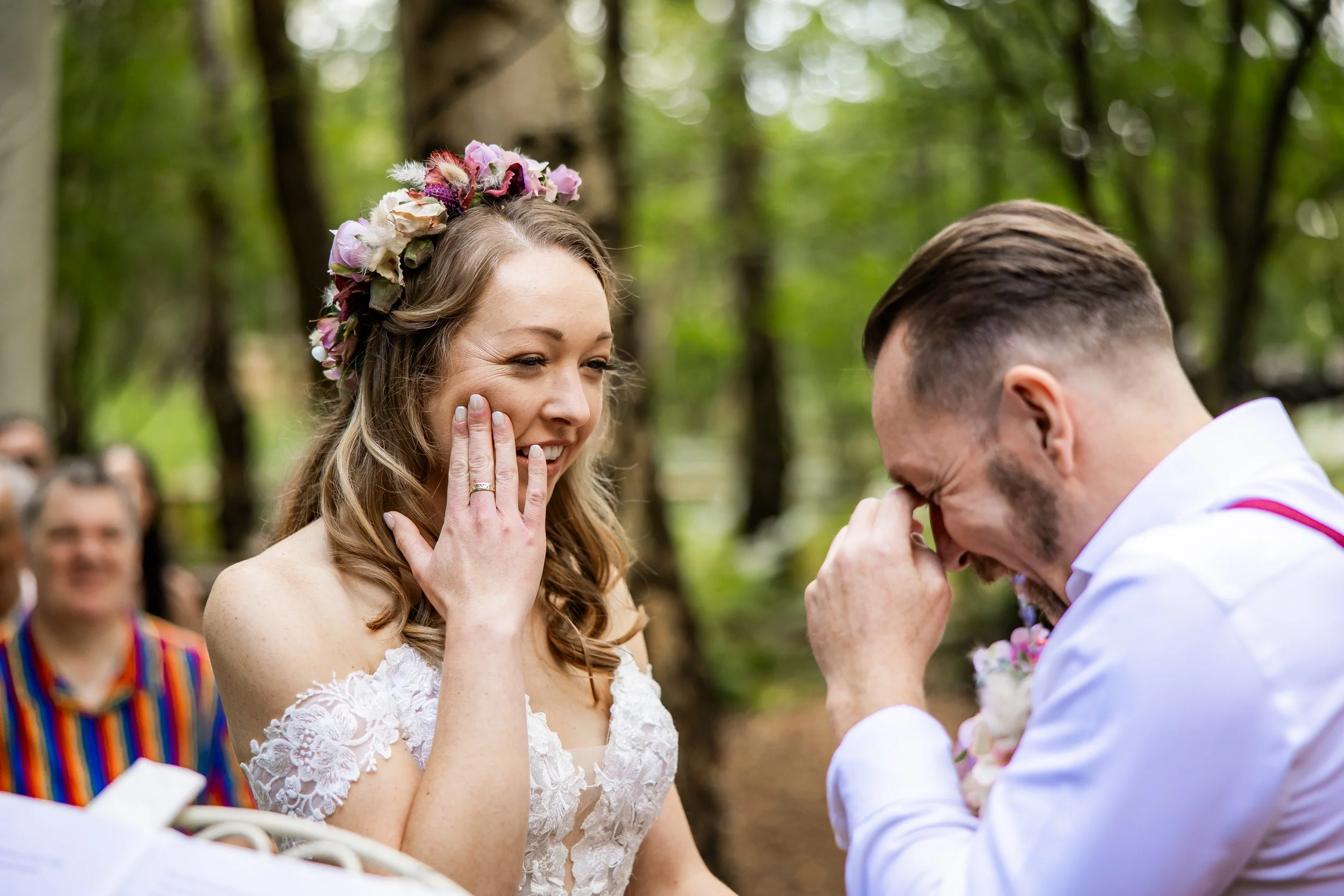 A woman with a floral crown and lace wedding dress is emotional, smiling with her hand near her cheek, as a man in a white shirt and boutonniere holds his face with his hand during a wedding ceremony in a forest.