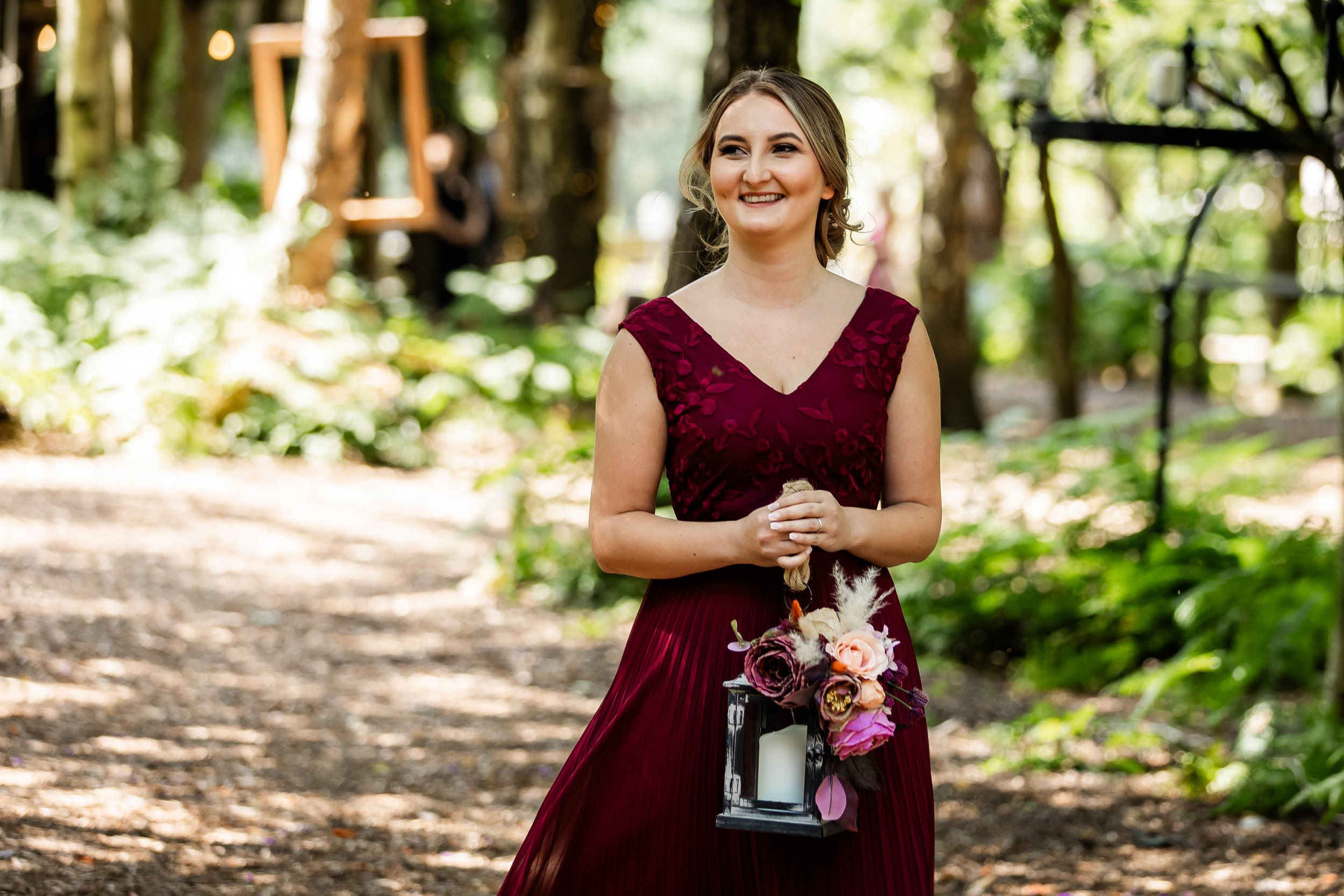 A smiling woman in a burgundy dress holding a lantern with a candle and a flower arrangement, standing in a wooded outdoor area.