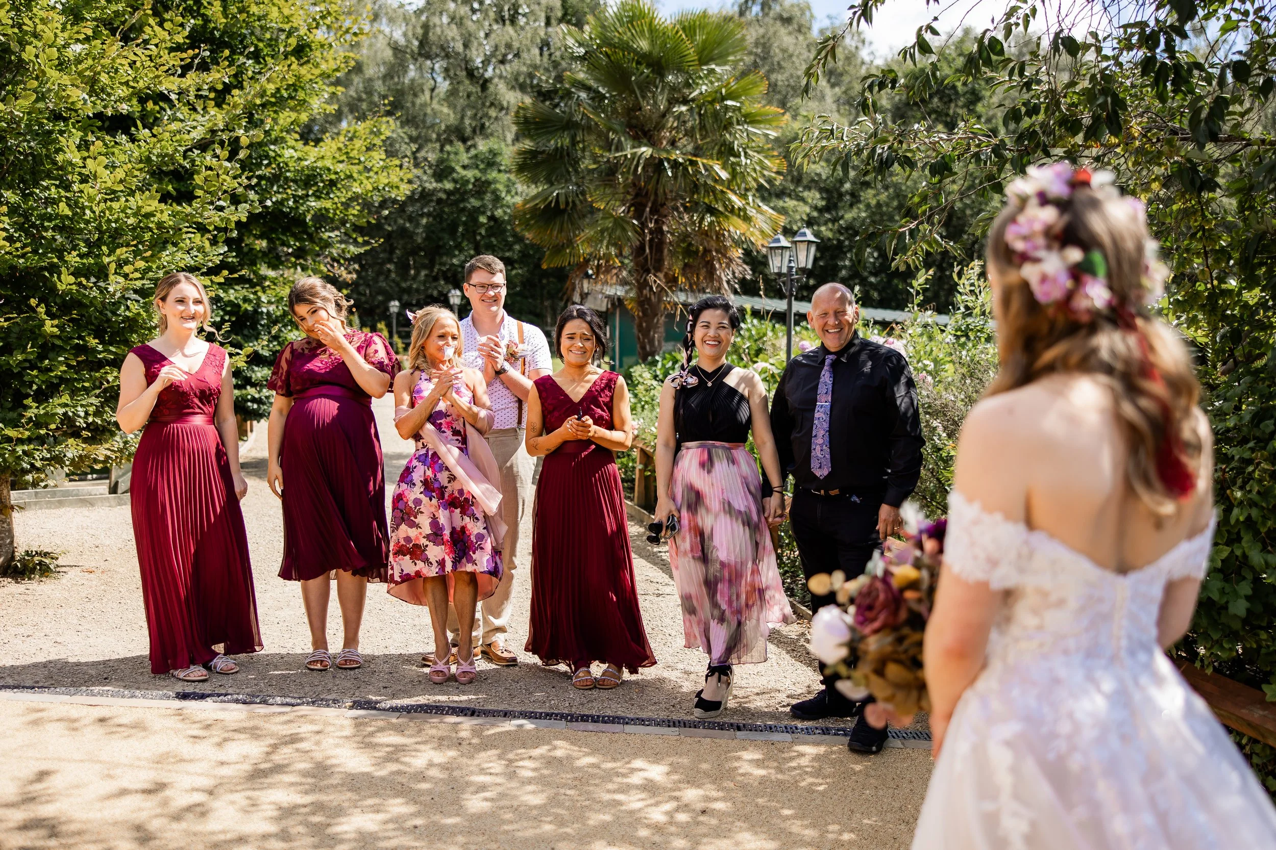 Bride with floral crown and off-shoulder wedding dress holding a bouquet, facing a group of women and men in colorful dresses and shirts, outdoors in a garden.