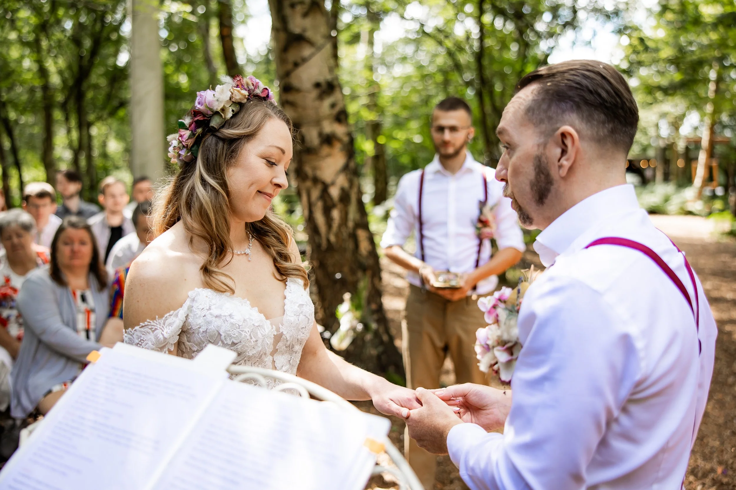 A bride and groom exchange rings during an outdoor wedding ceremony in a forest with guests in the background.