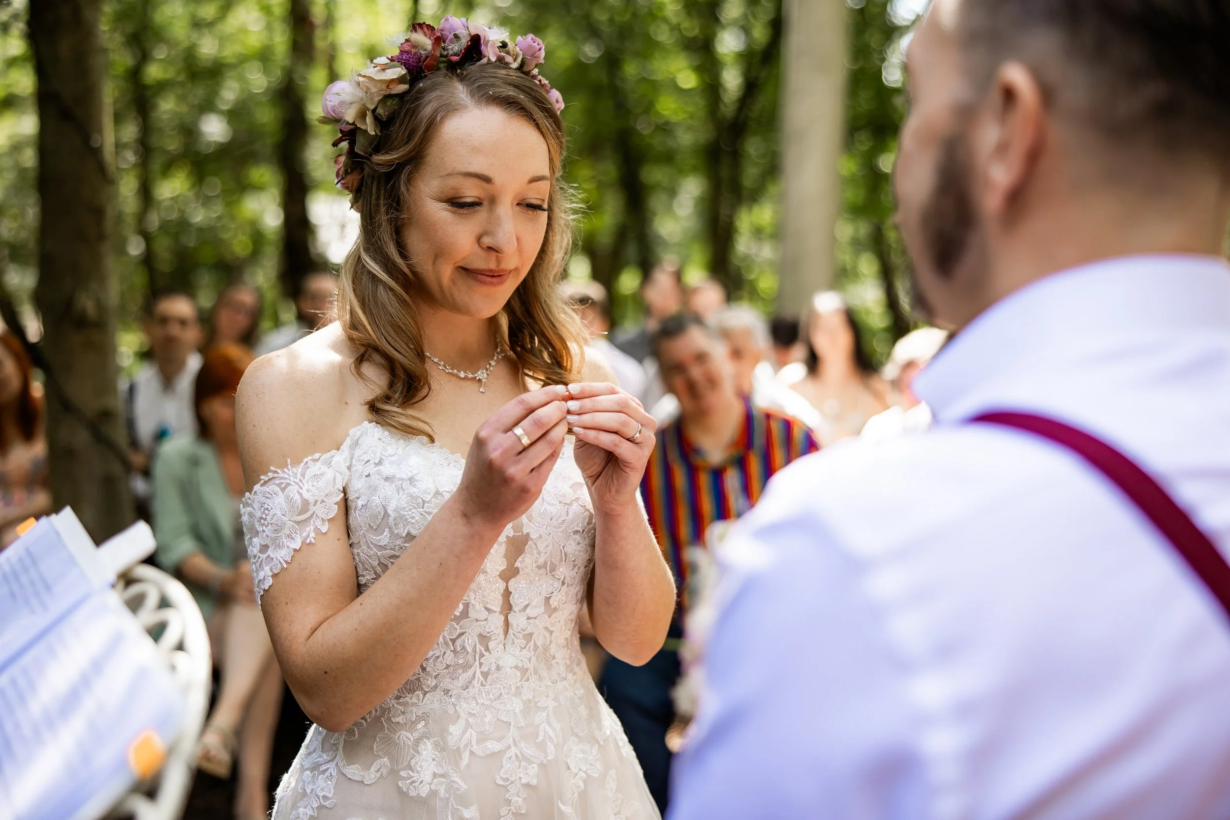 A woman with a floral crown and lace wedding dress holding a ring, looking down with a smile, during an outdoor wedding ceremony in a wooded area, with a man partially visible in the foreground and guests in the background.