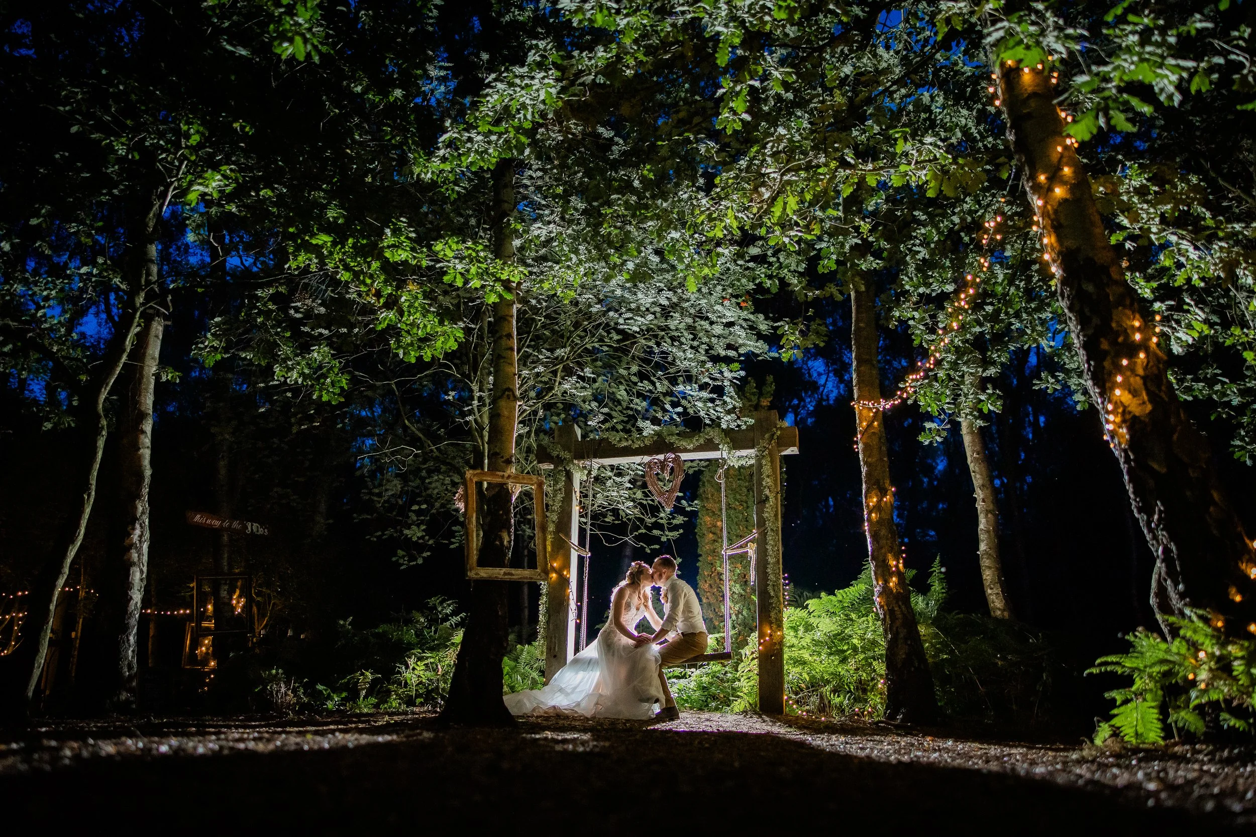 A newlywed couple sitting on a swing in a wooded area at night, surrounded by string lights and illuminated greenery, sharing a moment together.