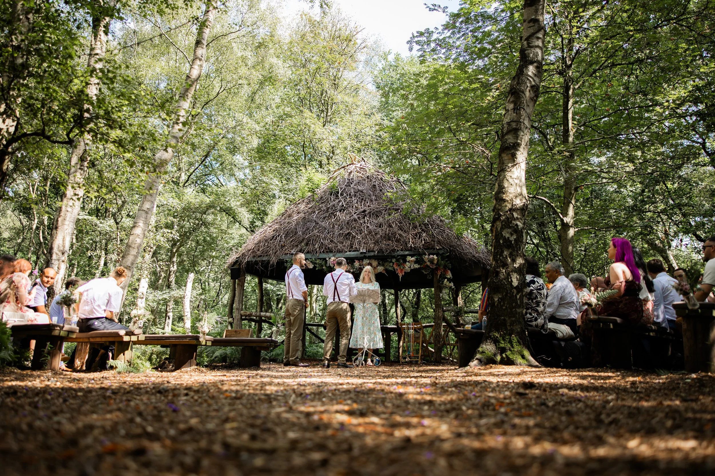 Outdoor wedding ceremony in a forest with people seated on benches and a couple standing under a thatched-roof gazebo.