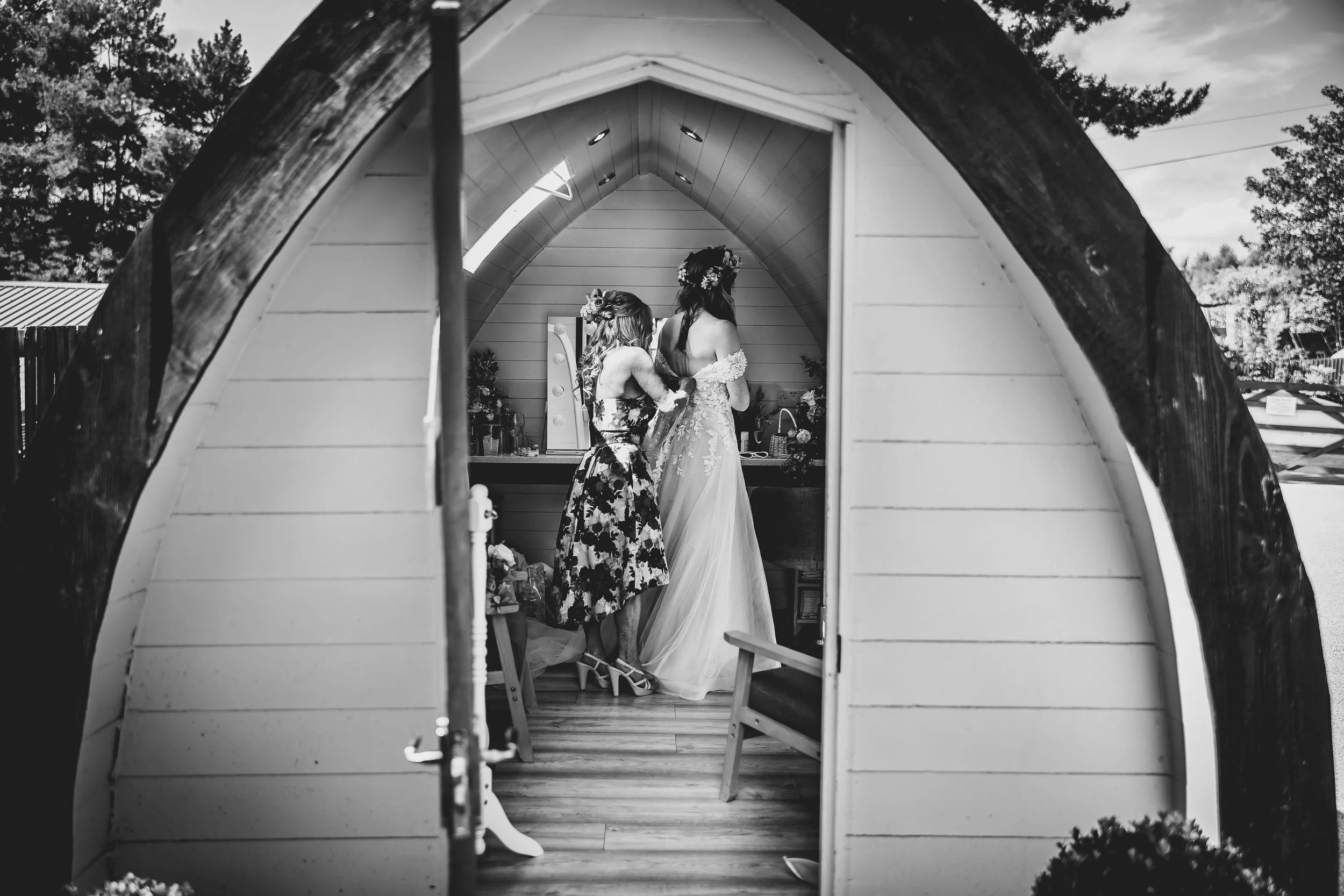 A woman in a wedding dress and a young girl with a floral dress and headband inside a small wooden cabin, fitting or preparing for a wedding.