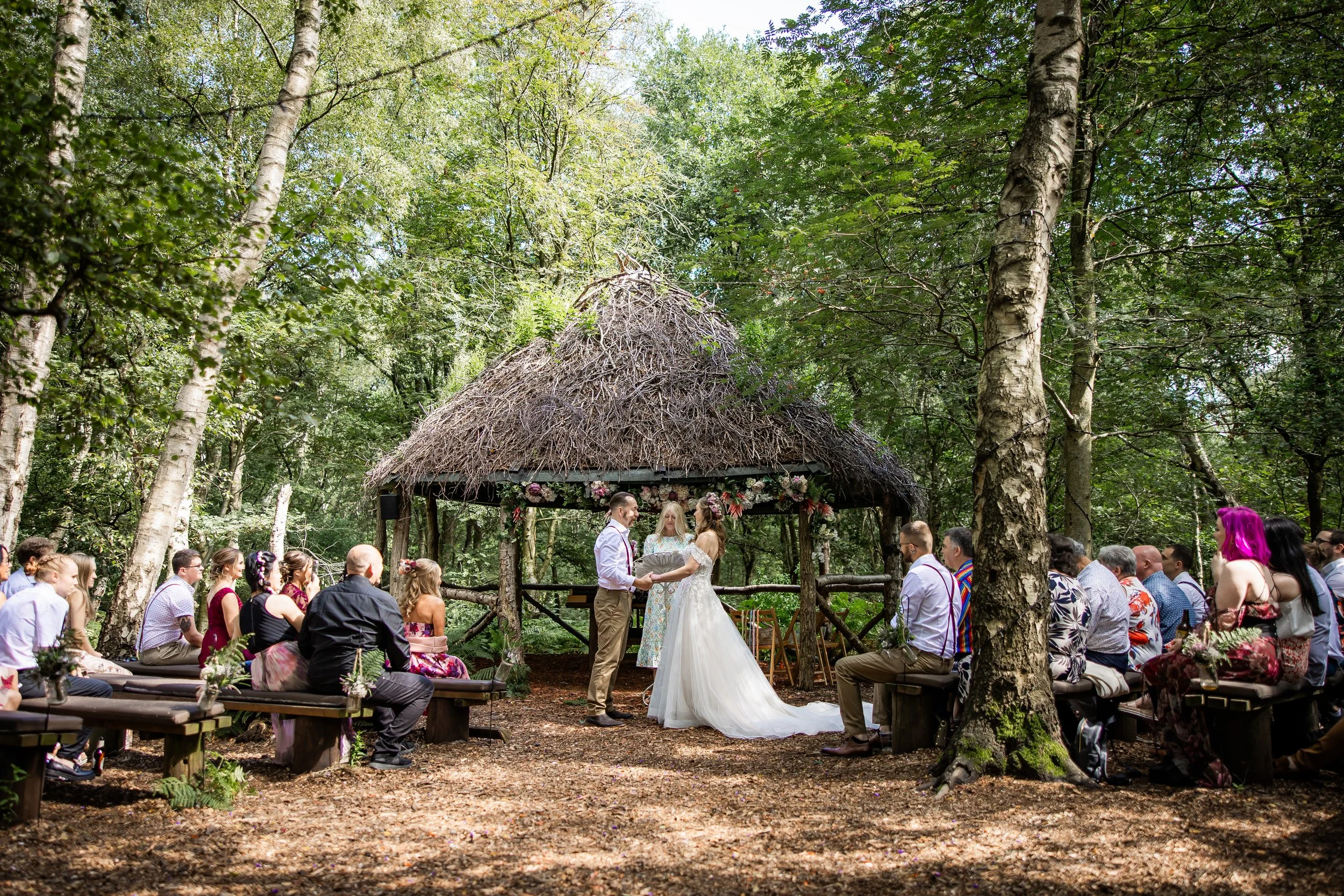 A wedding ceremony taking place outdoors in a forest clearing, with a thatched-roof gazebo decorated with flowers. The bride and groom are exchanging vows, surrounded by seated guests on benches at Cheshire Woodland Weddings.