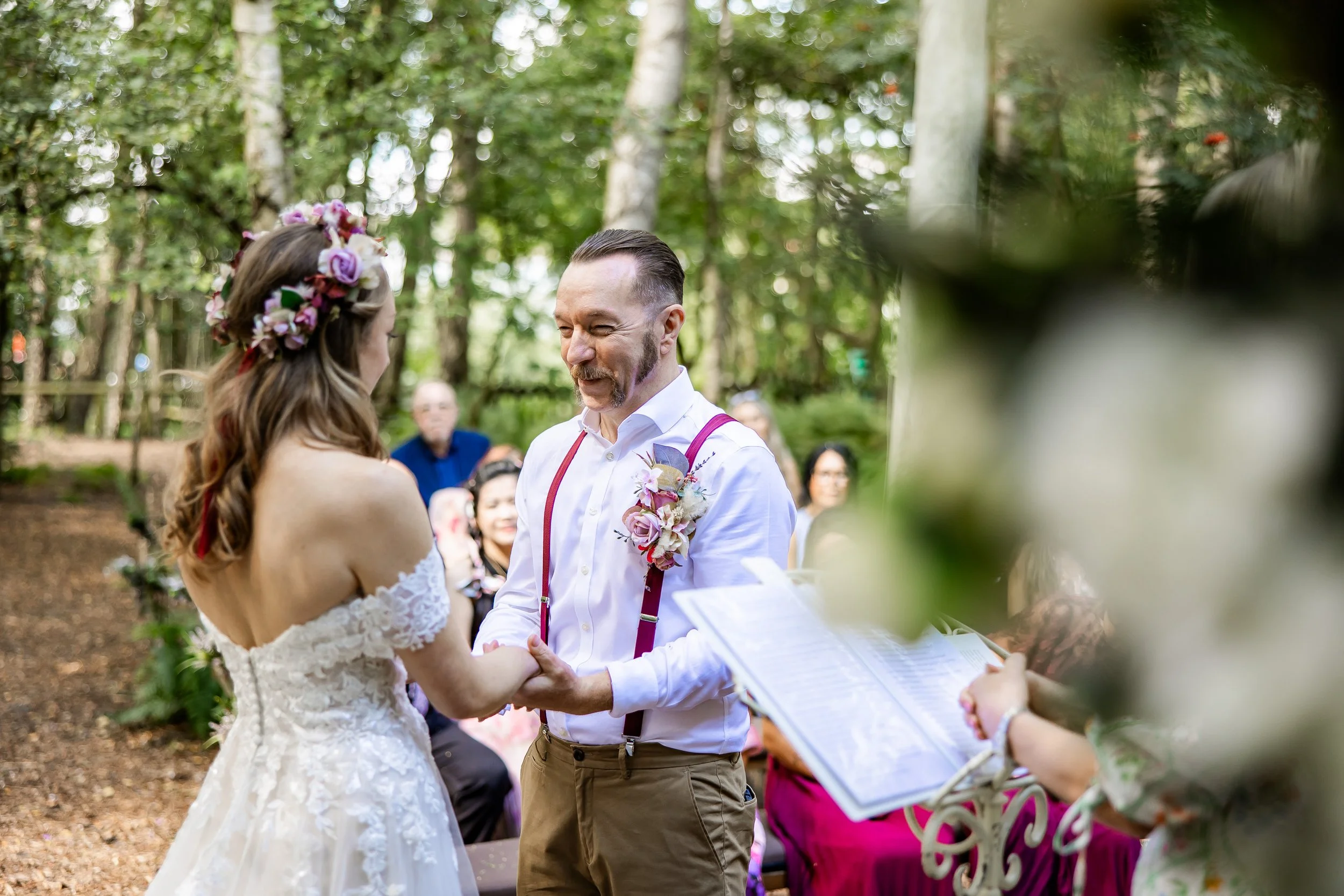 A couple getting married outdoors, holding hands, with guests in the background, surrounded by trees.