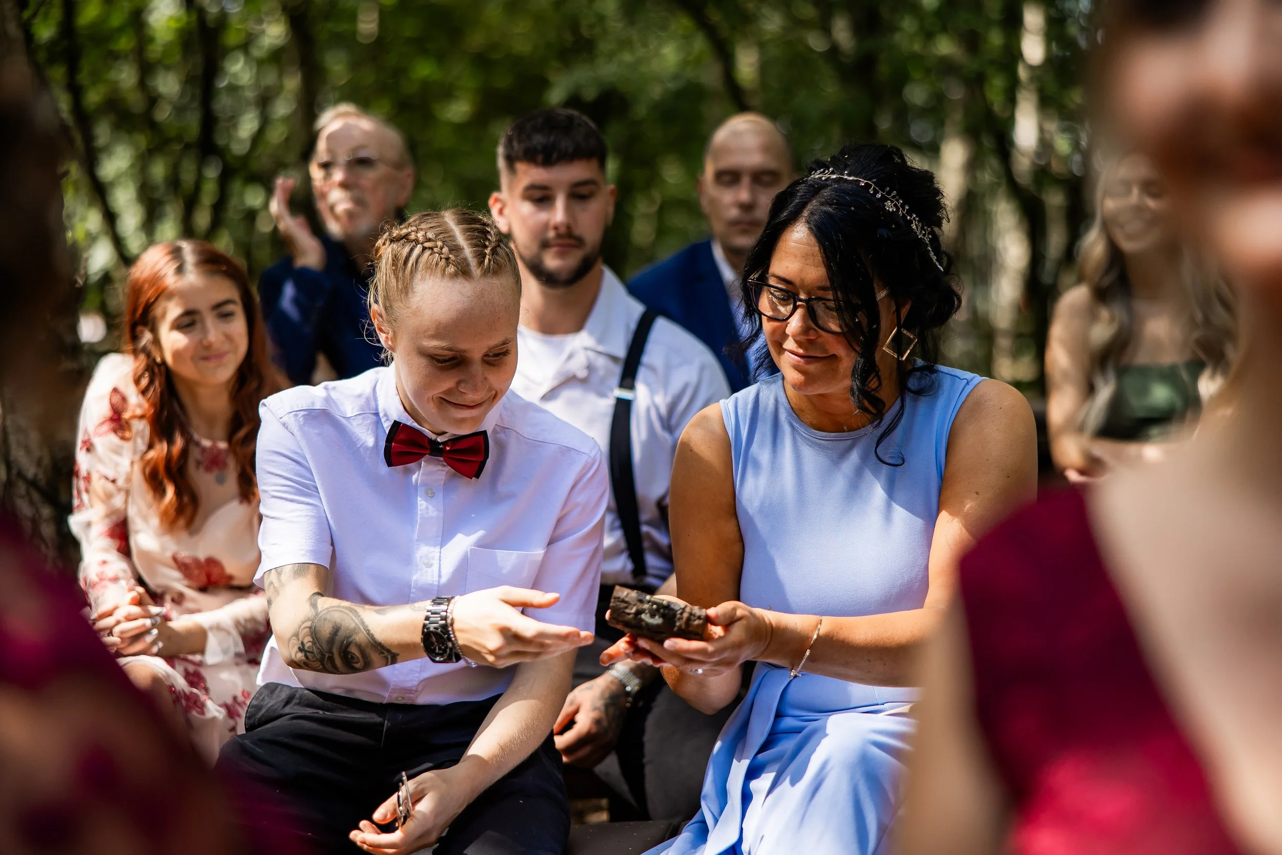 Group of people outdoors, with woman and young person exchanging a small gift, others watching, surrounded by trees.