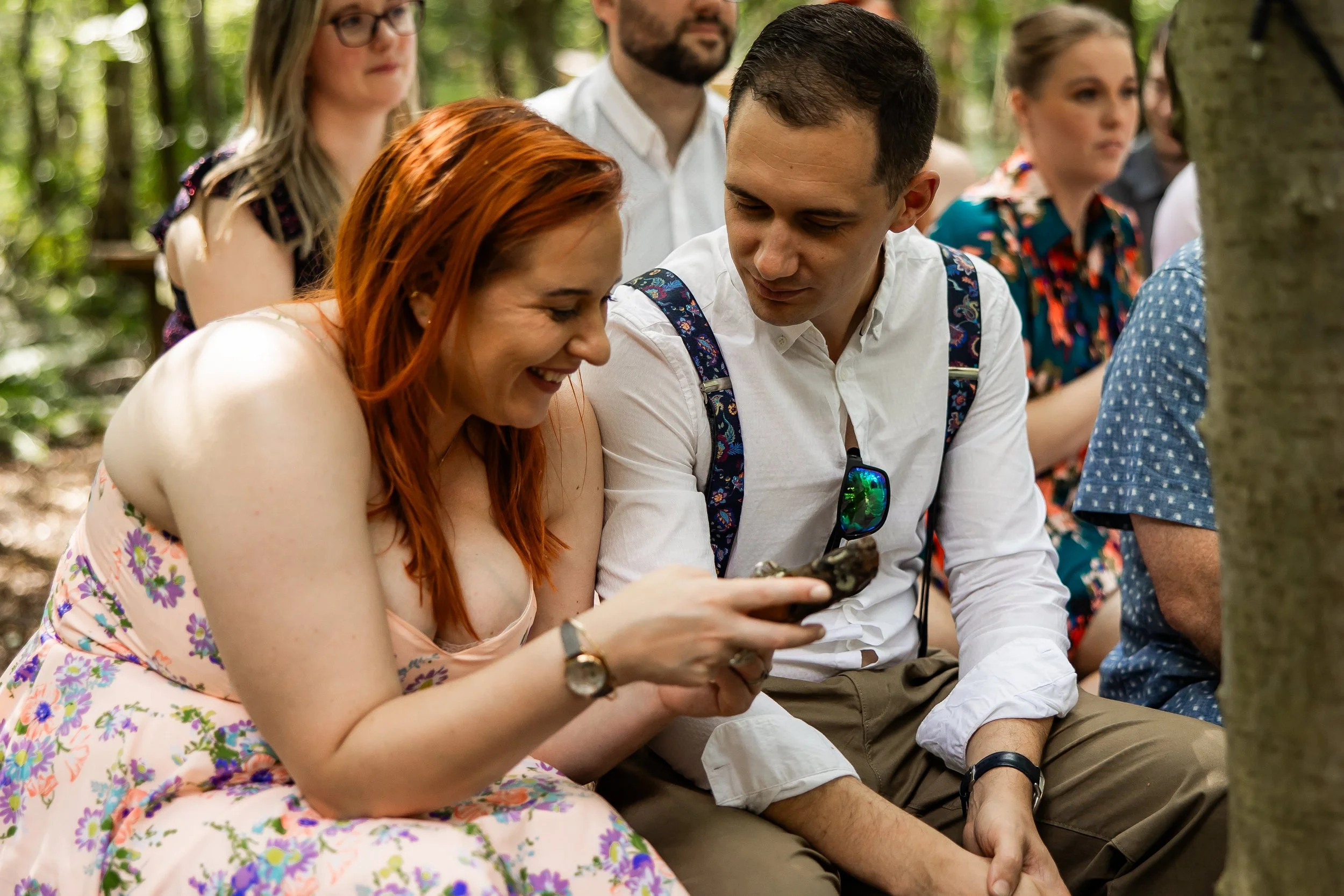 Group of people sitting outdoors in a forest, two young adults in the foreground sharing a laugh while looking at a phone, others in the background engaged in conversation.