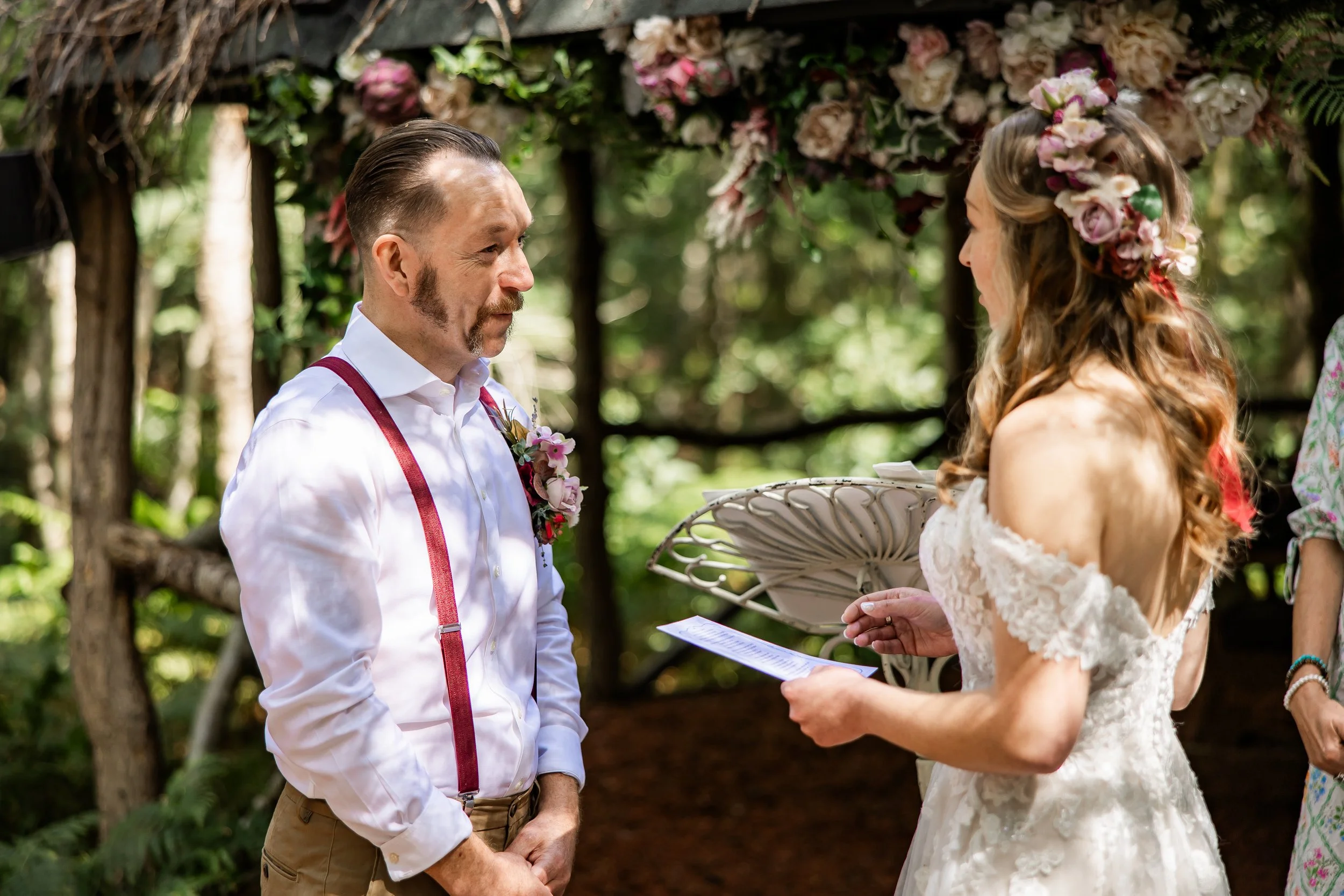 A bride and groom exchange vows during a wedding ceremony in a wooded outdoor setting. The groom wears a white shirt with red suspenders and floral boutonniere, while the bride is in a lace off-the-shoulder dress with a floral crown. The bride holds 