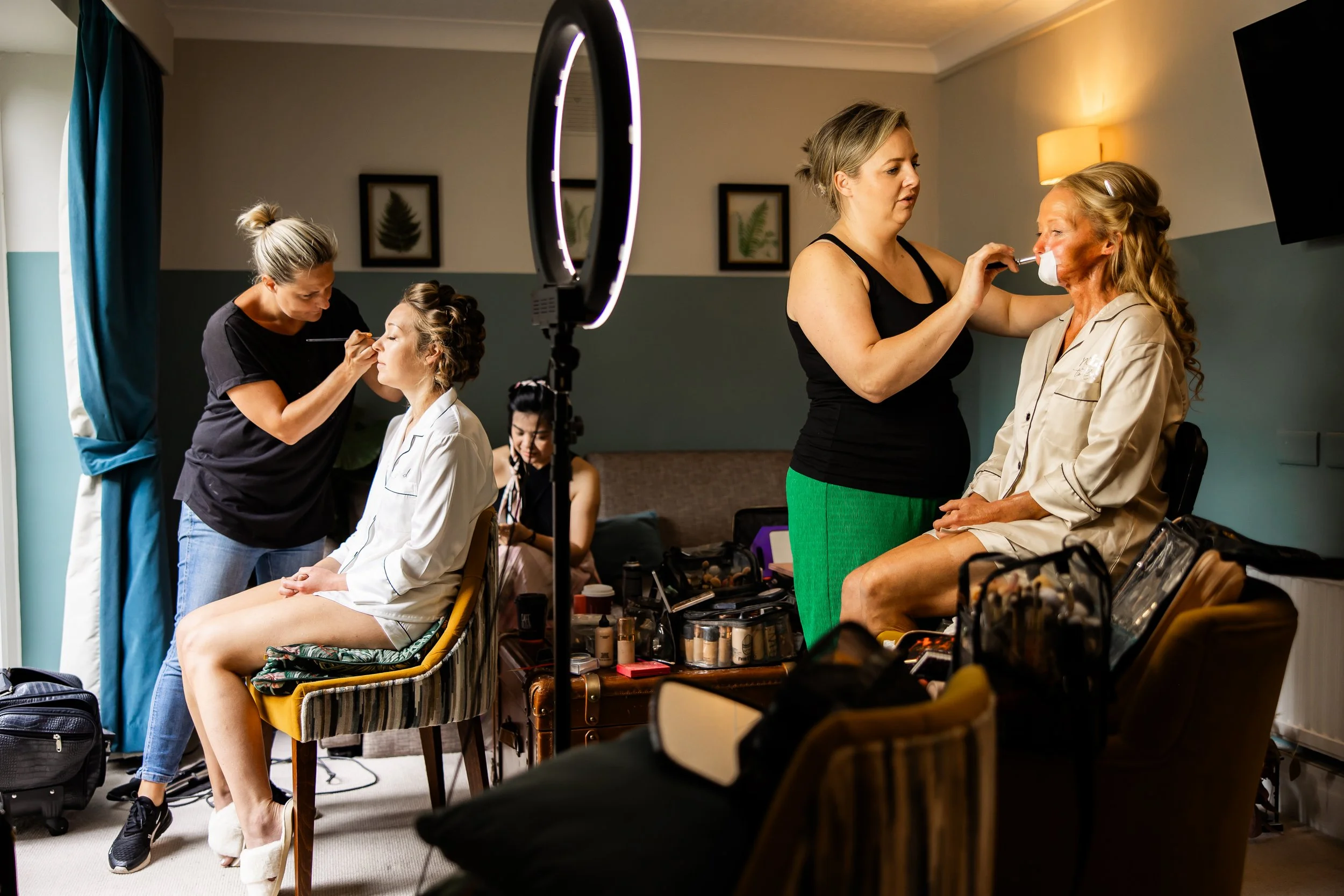 Two women applying makeup on a seated woman in a beige robe, with makeup artists working and makeup supplies around in a room with a ring light and framed botanical artwork.