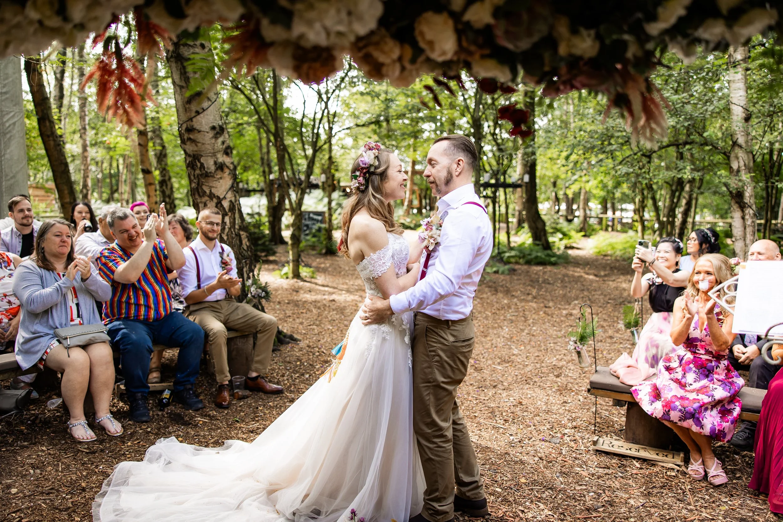 A couple shares a first dance at an outdoor wedding ceremony in a forested area, with guests seated on benches clapping and taking photos.