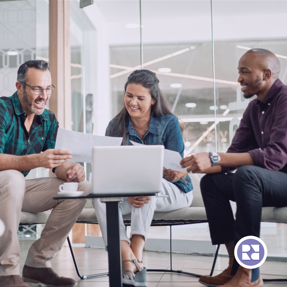 two men and a woman sitting around a computer smiling and sharing paper reports with each other