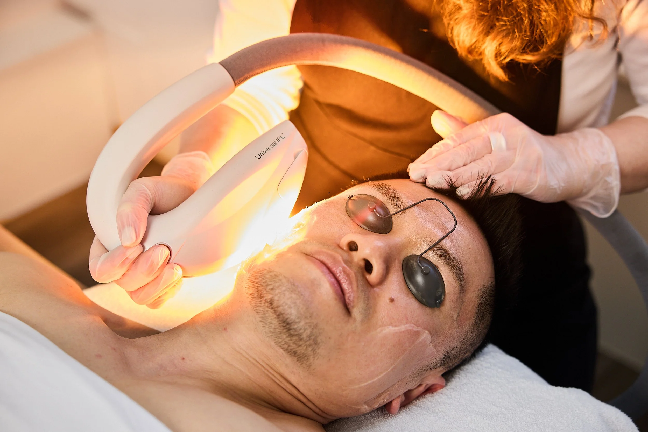 A man lying down with eyes closed, undergoing a medical treatment that involves a device emitting warm light. A healthcare professional, wearing gloves, is operating the device positioned above his face.