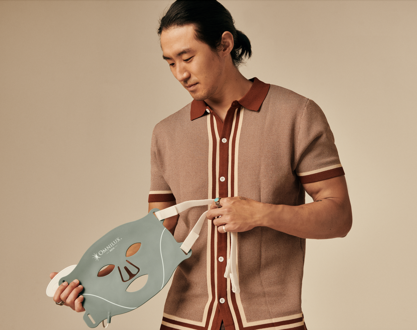 A man with dark hair tied back, wearing a retro brown, beige, and red striped shirt, is holding a grey face mask with cut-outs for eyes, nose, and mouth.