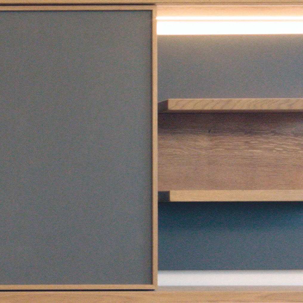 Close up image of a large custom made television cabinet, made from oak and grey panels. There is integrated lighting under the shelving and the tv is covered with sliding doors.