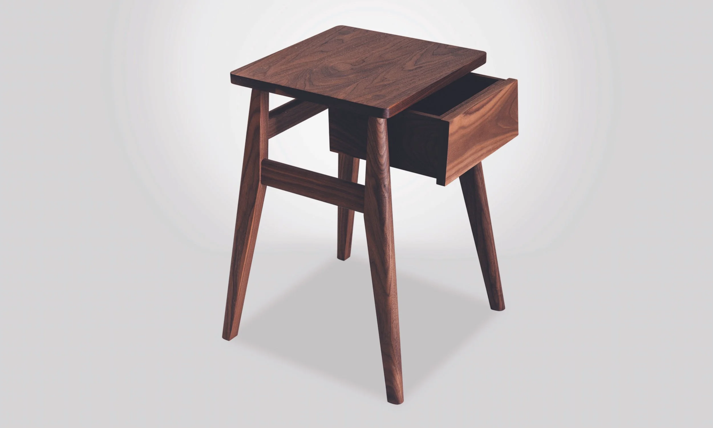 A wooden bedside table with a drawer. Made in solid walnut wood, the table has a smooth finish and eye catching detailed joinery. The level of quality only achieved by skilled crafts people.