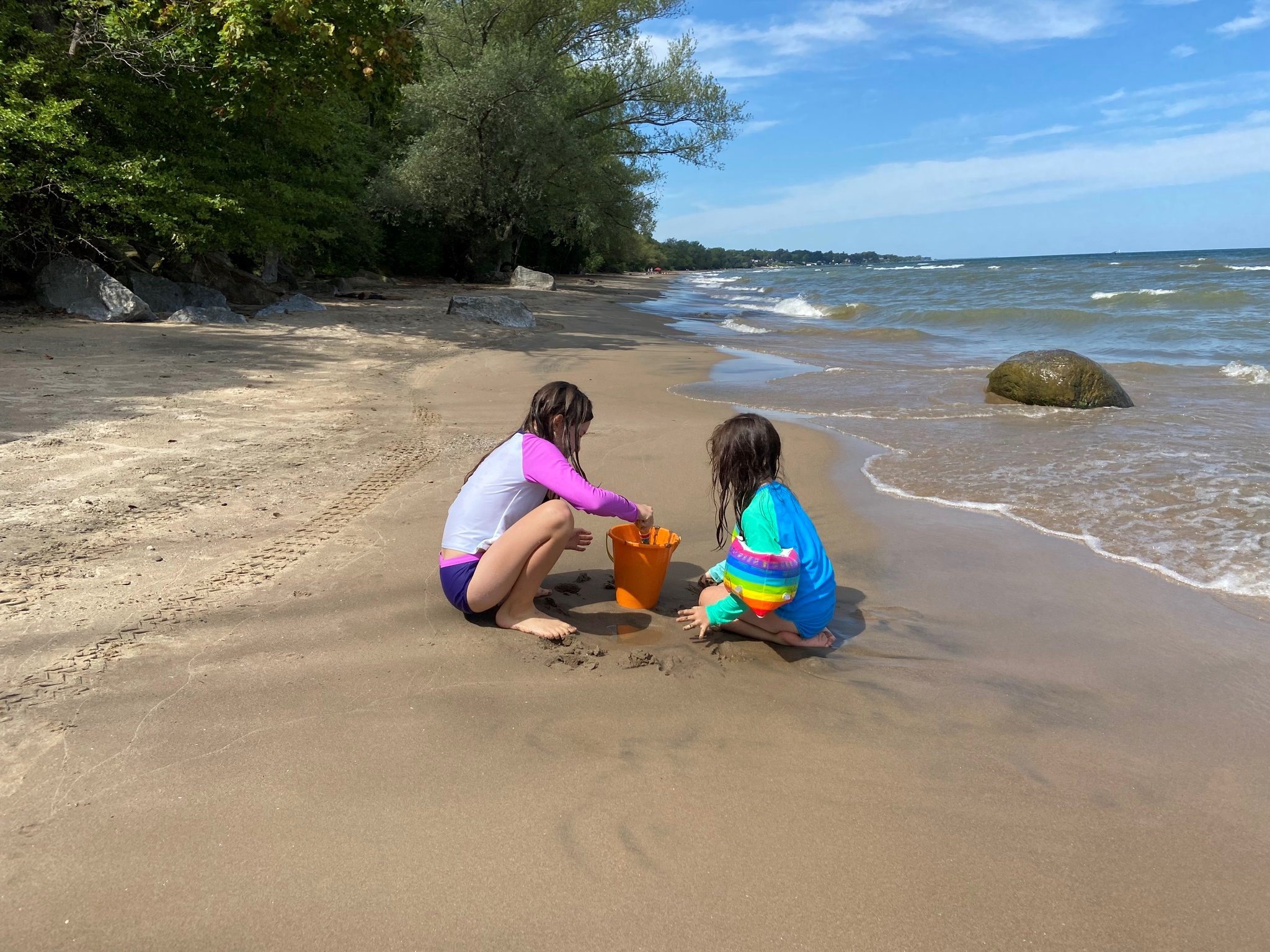 Two children in swimsuits and rainbow arm floaties play in the sand at a Lake Ontario beach. The sky is blue and green trees and large rocks line the shore.