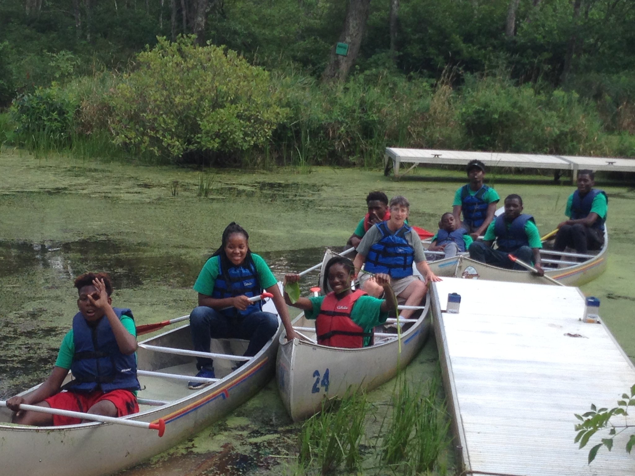 9 teens wear life jackets and sit in 4 canoes near a dock in Sodus Bay.
