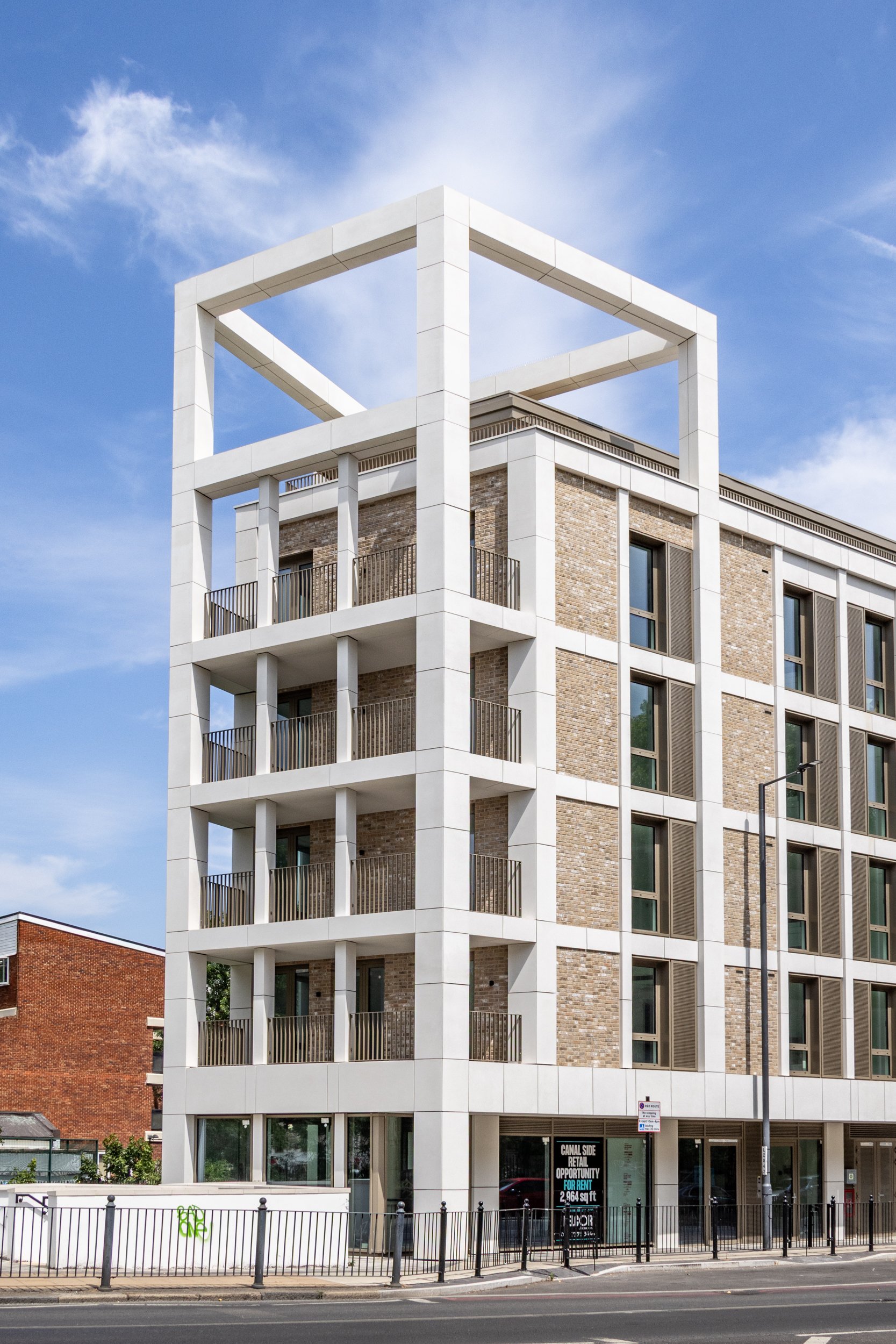 Modern multi-story building with a white geometric frame on top, brick and glass facade, street view with sidewalk and metal railings, clear blue sky.