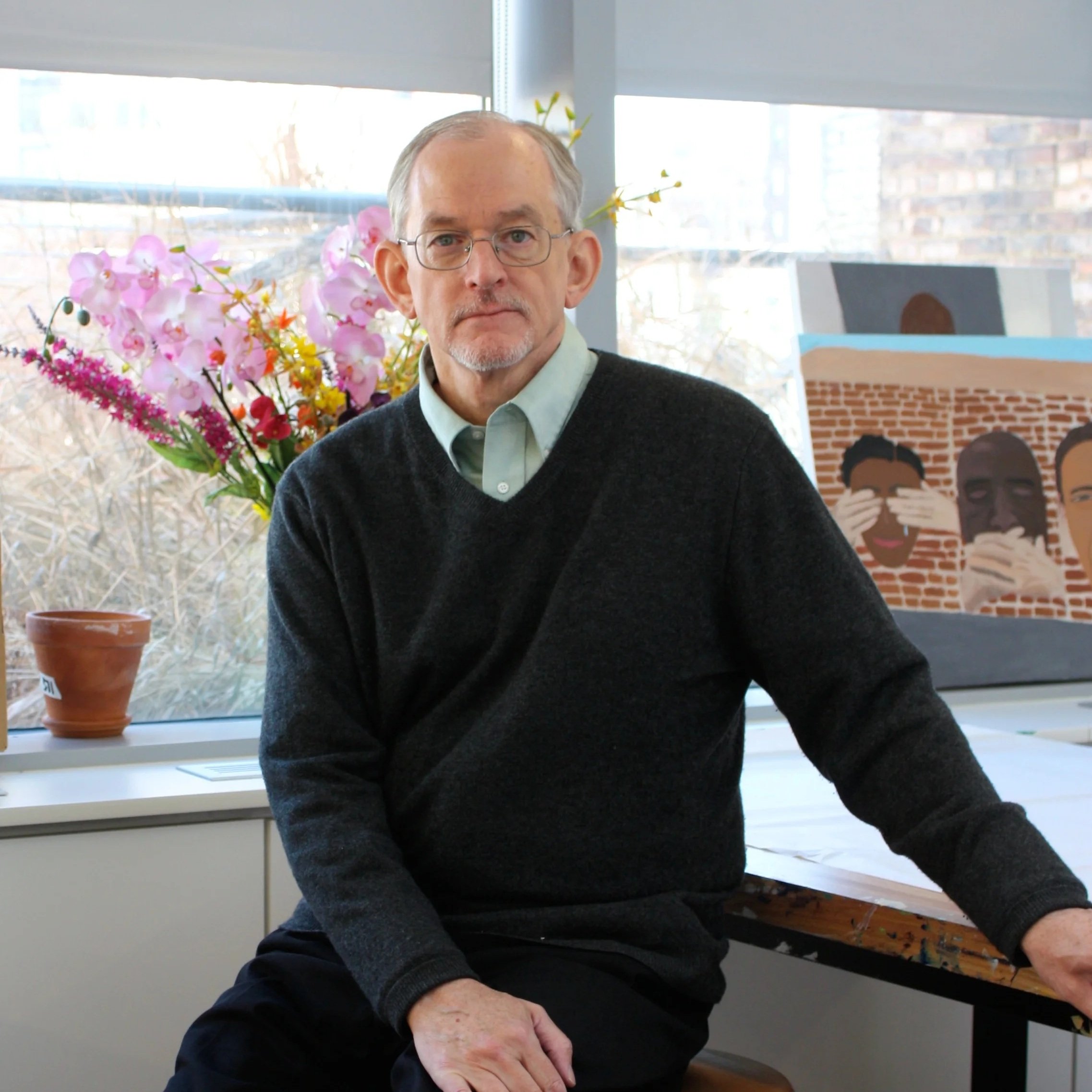 John Loughery seated in a sunlit studio in a charcoal V-neck sweater, with orchids and a painting behind him