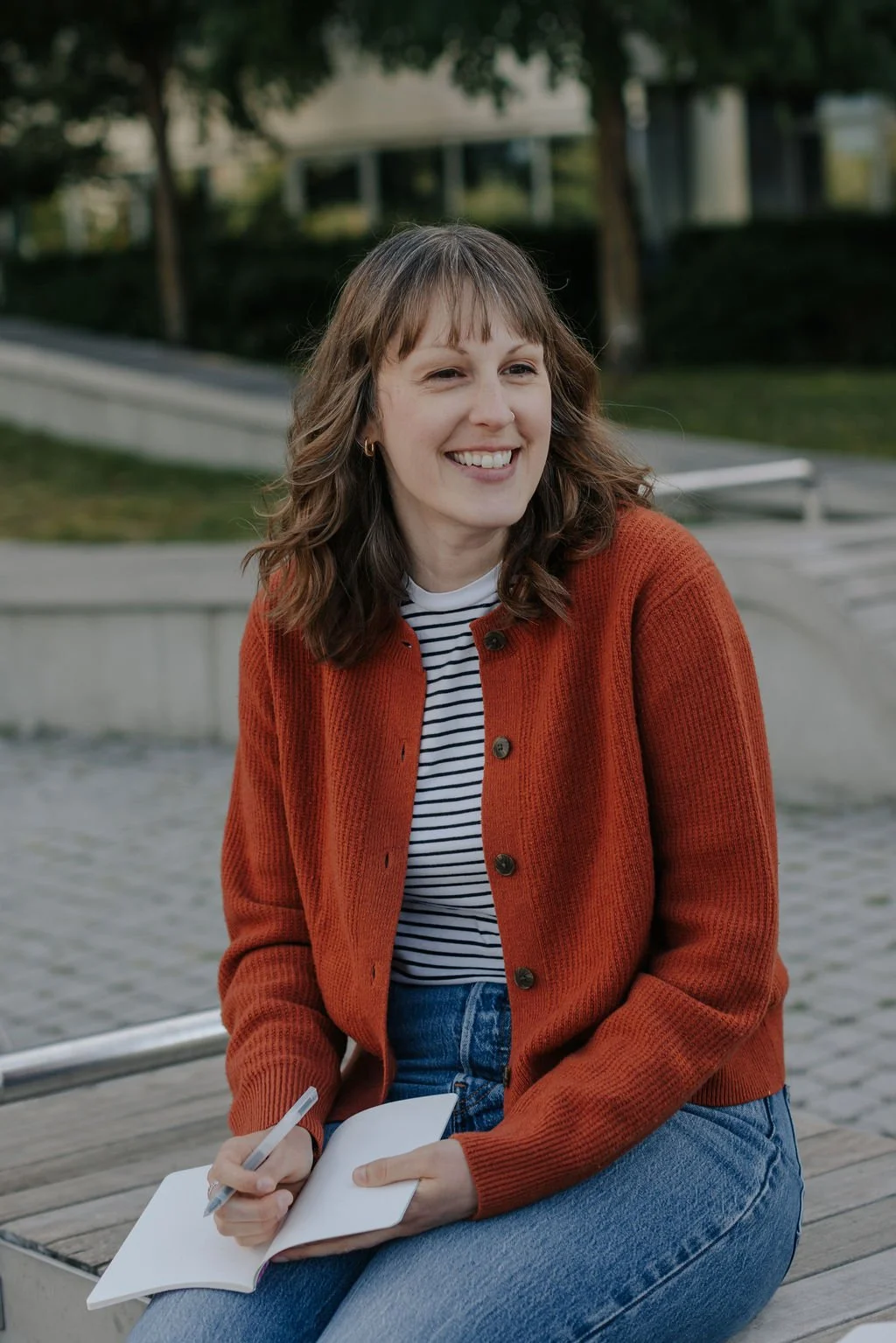 Emily, a white woman with long hair wearing an orange cardigan, smiles white holding a notebook