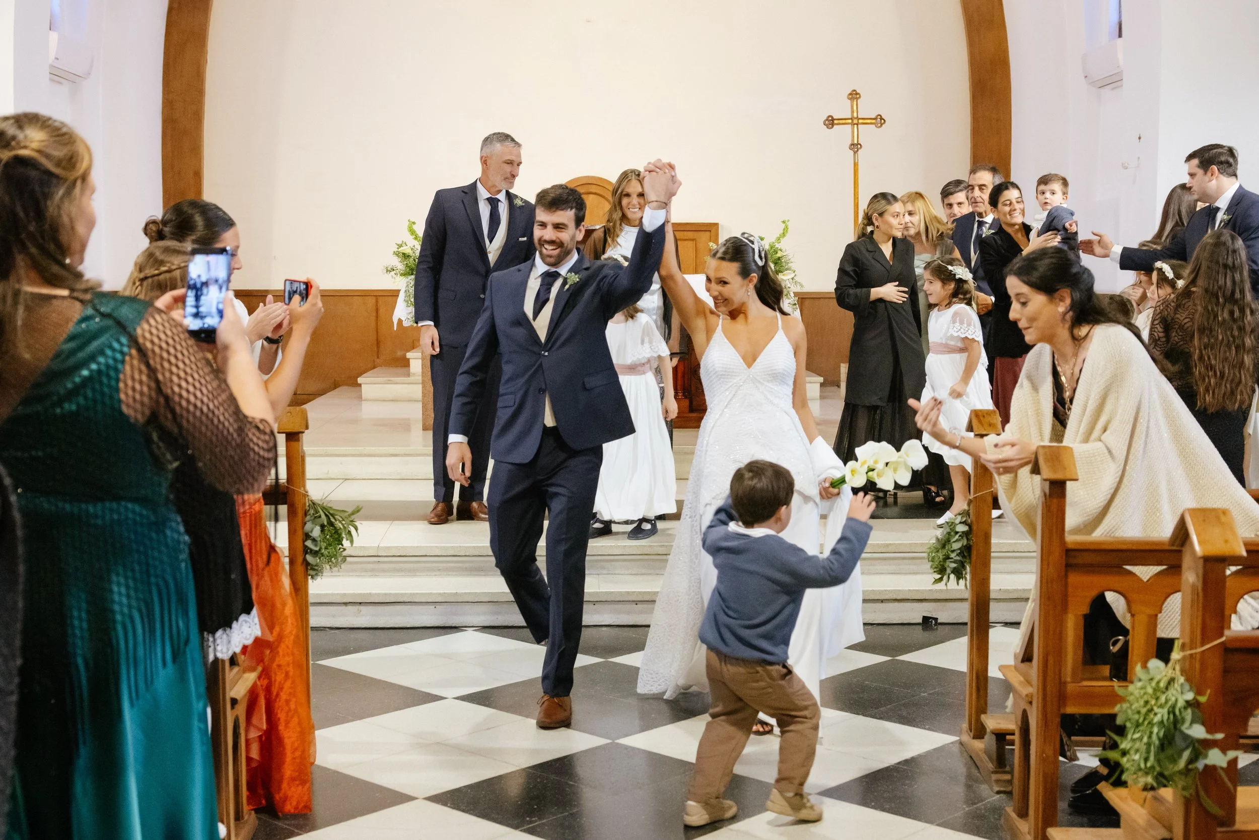 Pareja de novios en la iglesia celebrando su boda, rodeados de invitados. La pareja se da la mano con sonrisas, mientras algunos invitados toman fotos y observan la ceremonia.