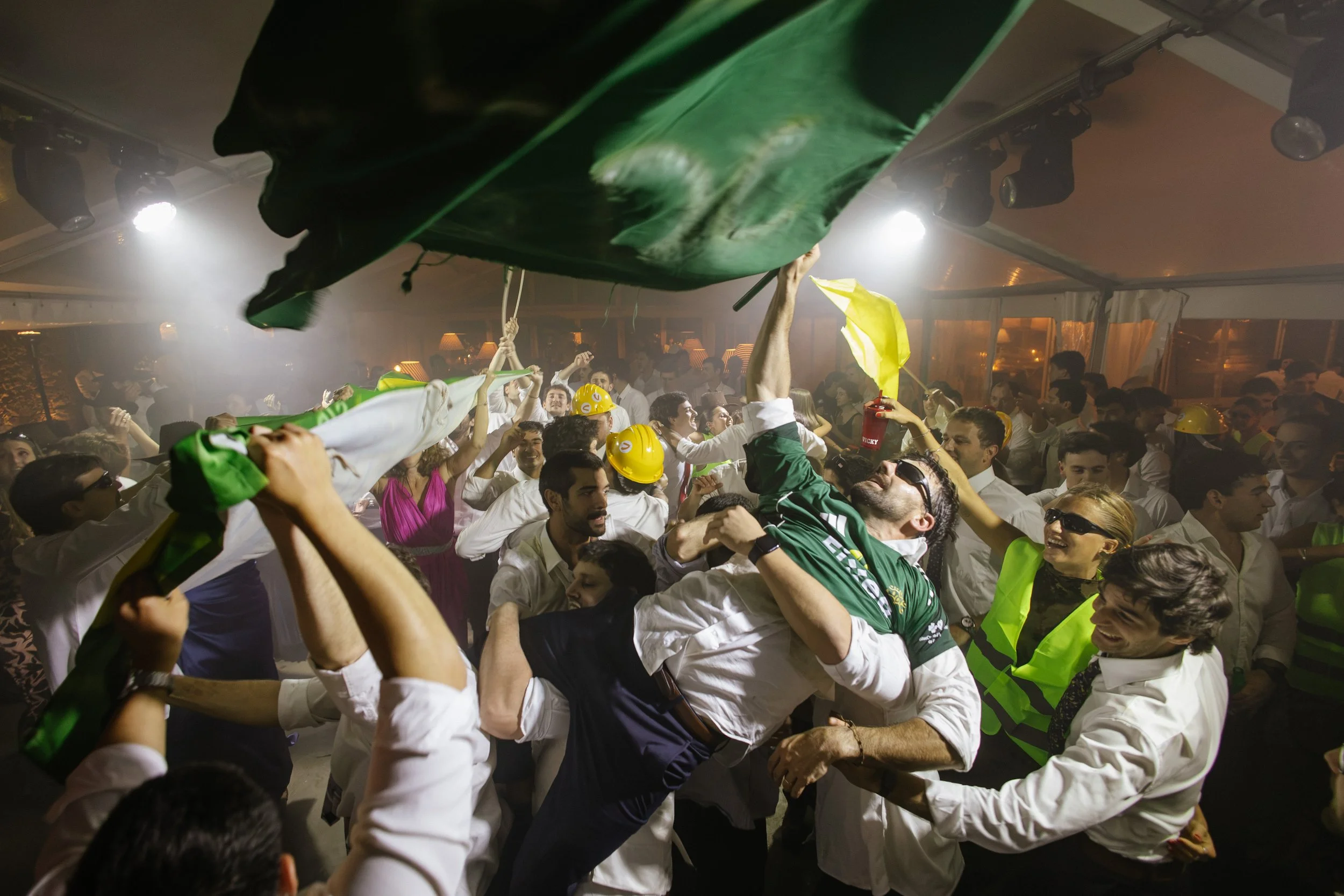Personas celebrando en una reunión, muchas sosteniendo banderas y usando cascos amarillos, en un ambiente festivo y enérgico.
