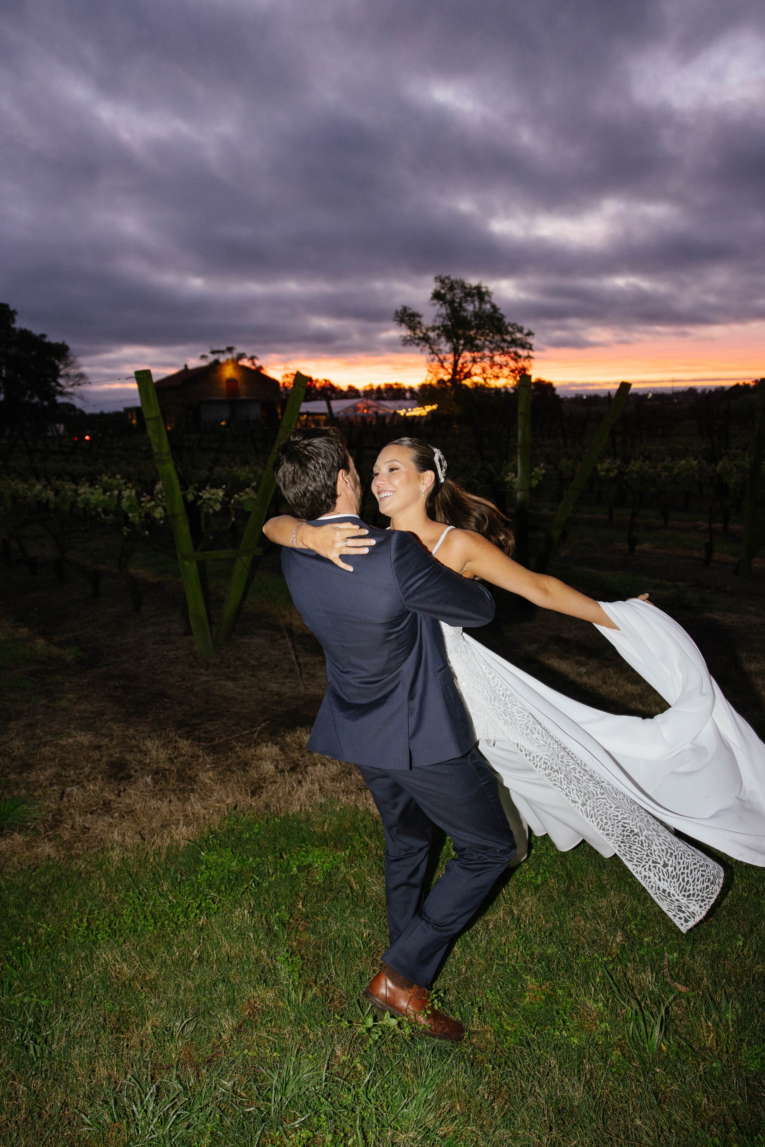 Pareja bailando en un campo al atardecer, la mujer lleva vestido blanco y el hombre un traje oscuro, fondo de cielo nublado y árboles.