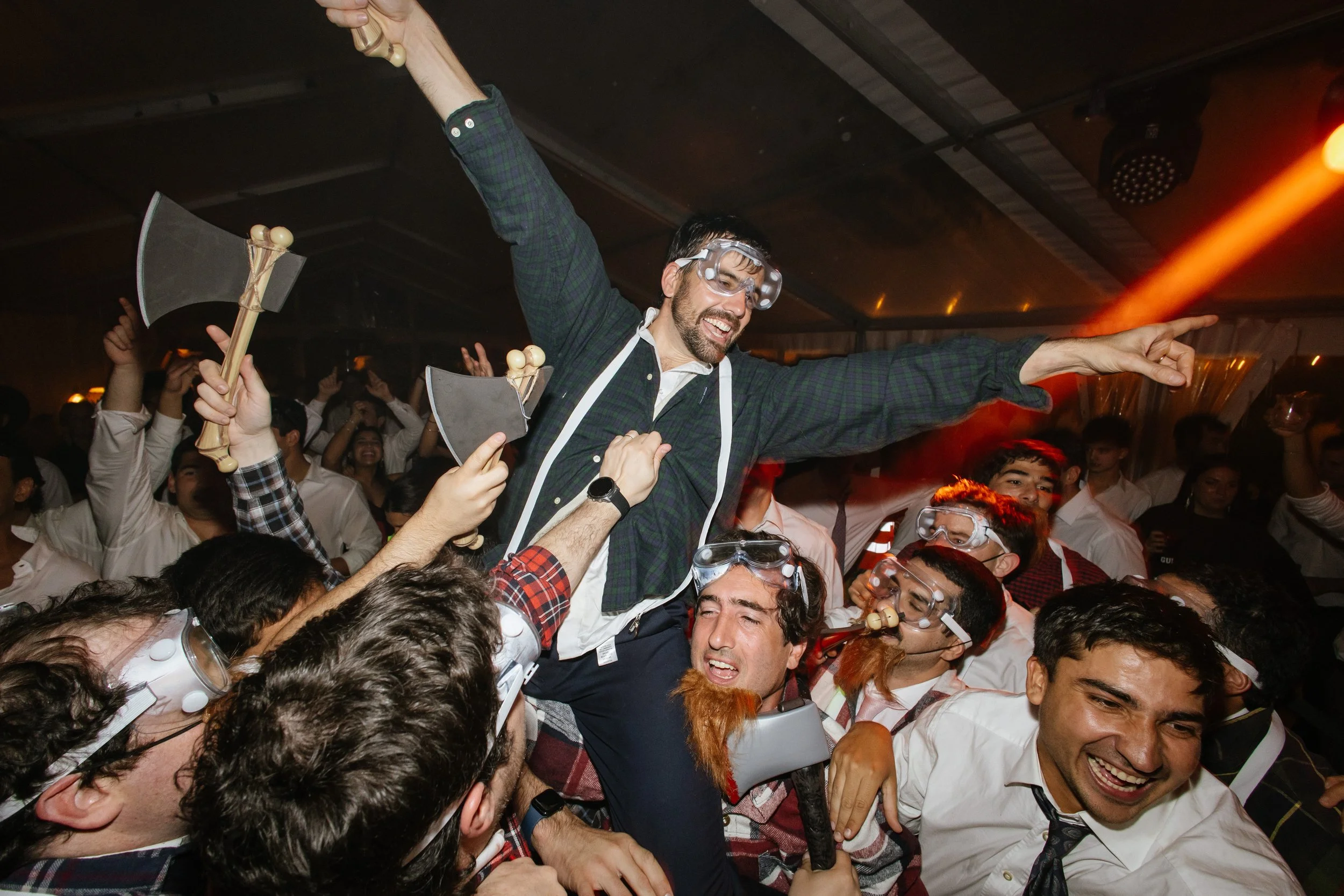 Persona levantándose en medio de una multitud en un evento, algunos con gafas de protección y sombreros, en ambiente festivo con luces rojas y oscuras.