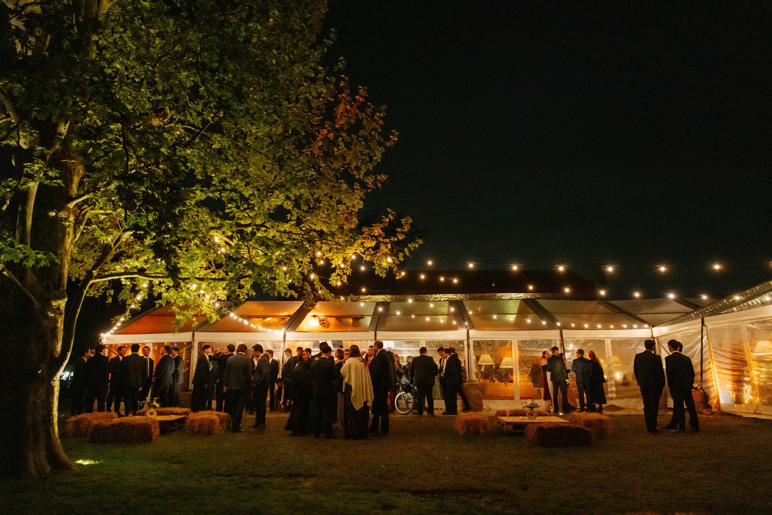 Un grupo de personas en un evento nocturno bajo una carpa transparente, con luces decorativas, en un jardín con árboles, asistencia en vestimenta formal.