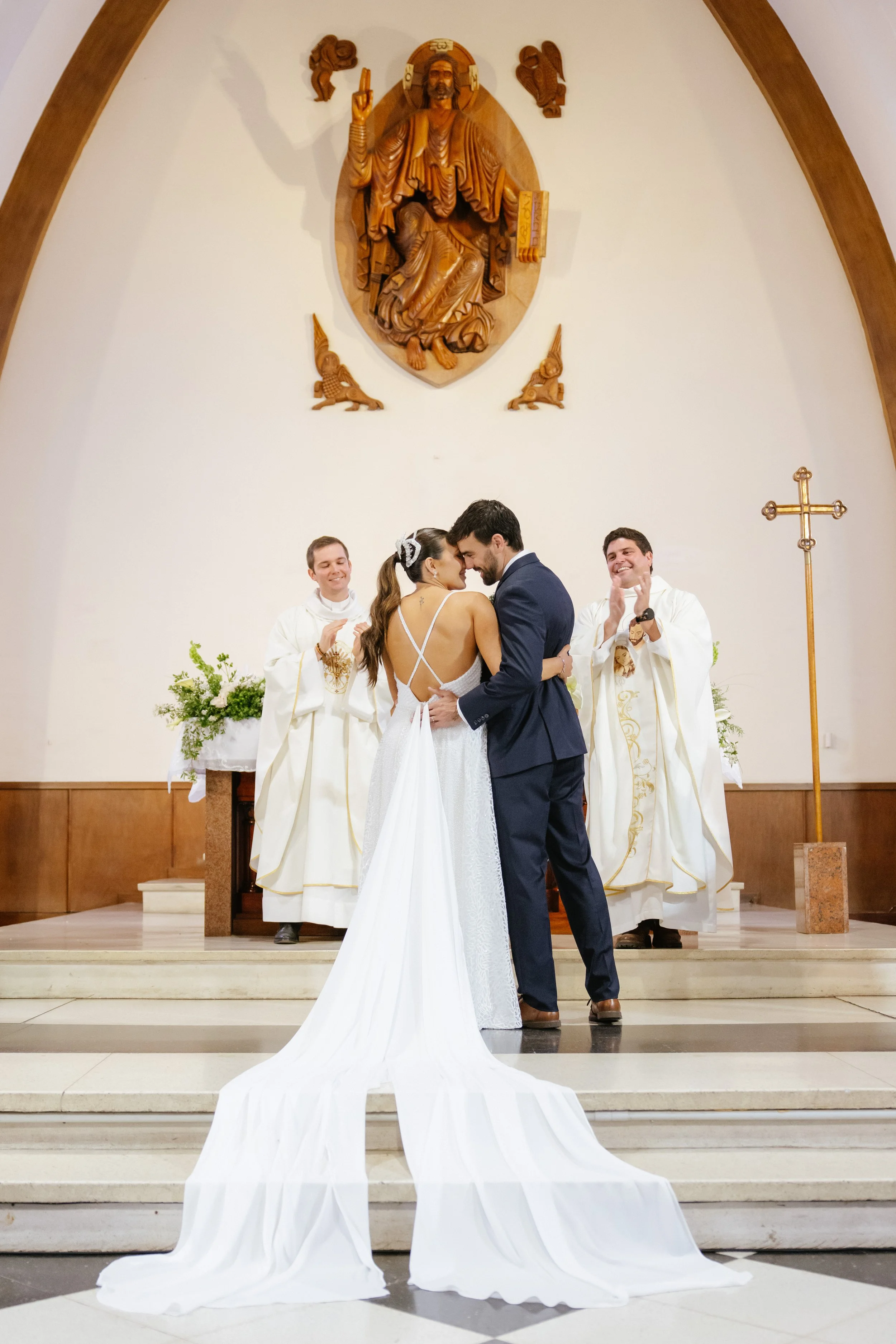 Una pareja de novios en el altar de una iglesia, con sacerdotes celebrando su boda, con un fondo de una obra de arte religiosa en madera
