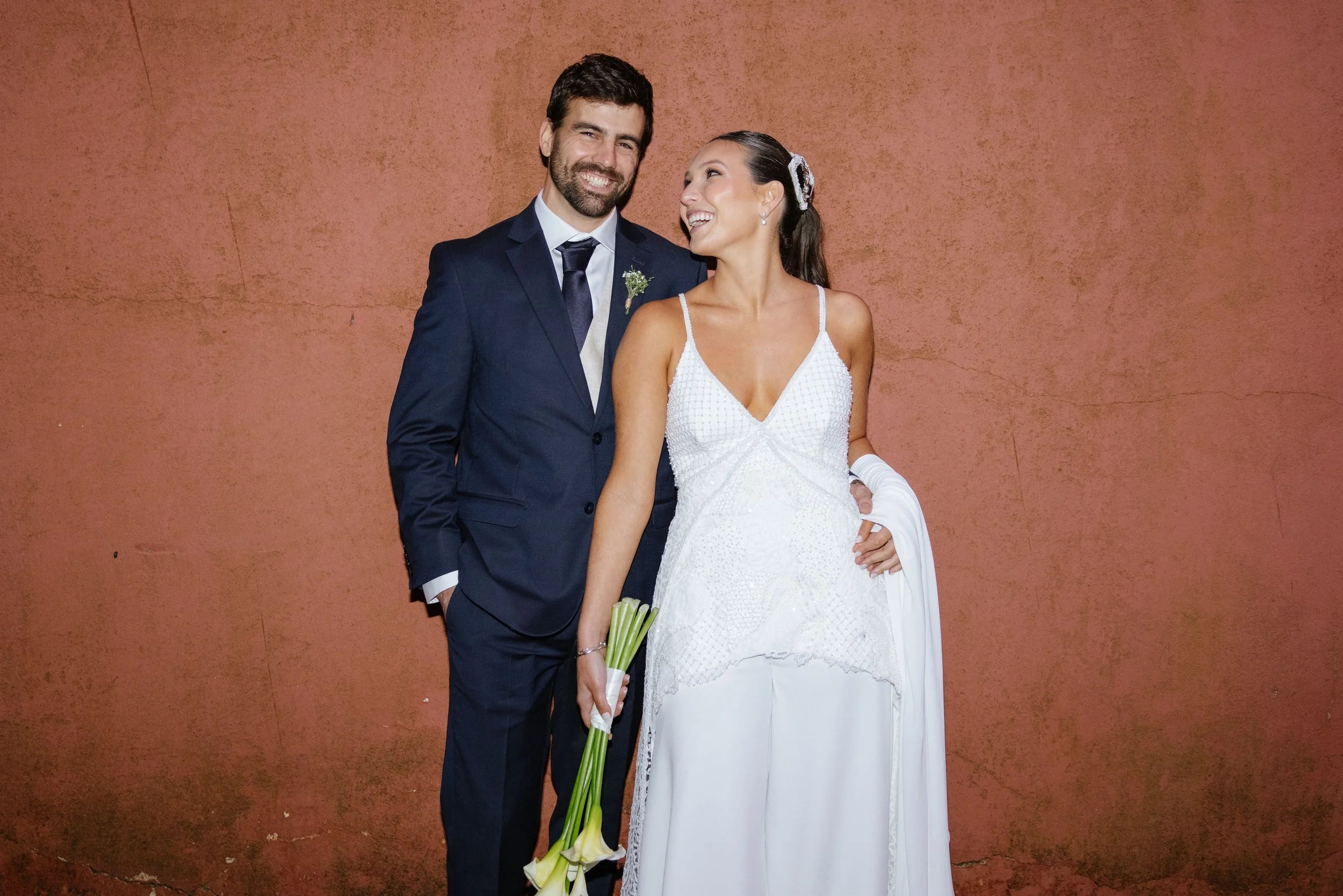 Pareja de luna de miel o recién casados posando juntos frente a una pared de color terracota. La mujer lleva un vestido blanco de boda y sostiene un ramo de flores blancas. Él viste un traje azul oscuro con corbata y camisa blanca, ambos lucen felice