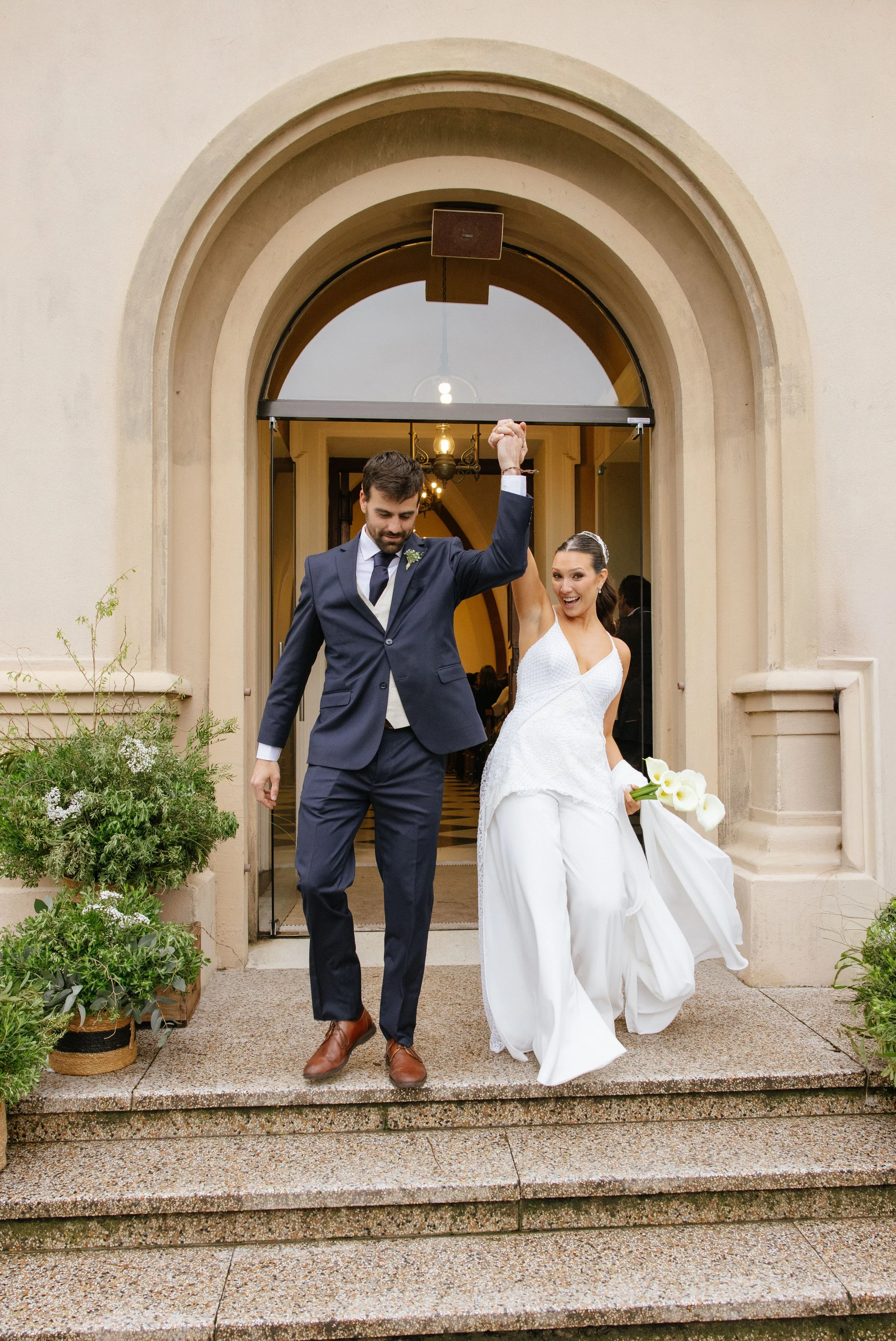 Una pareja de recién casados sale del edificio, la mujer lleva un vestido blanco y flores en las manos, y el hombre lleva un traje azul oscuro. Ambos parecen felices, con la mujer sonriendo y levantando la mano de su pareja en señal de victoria.