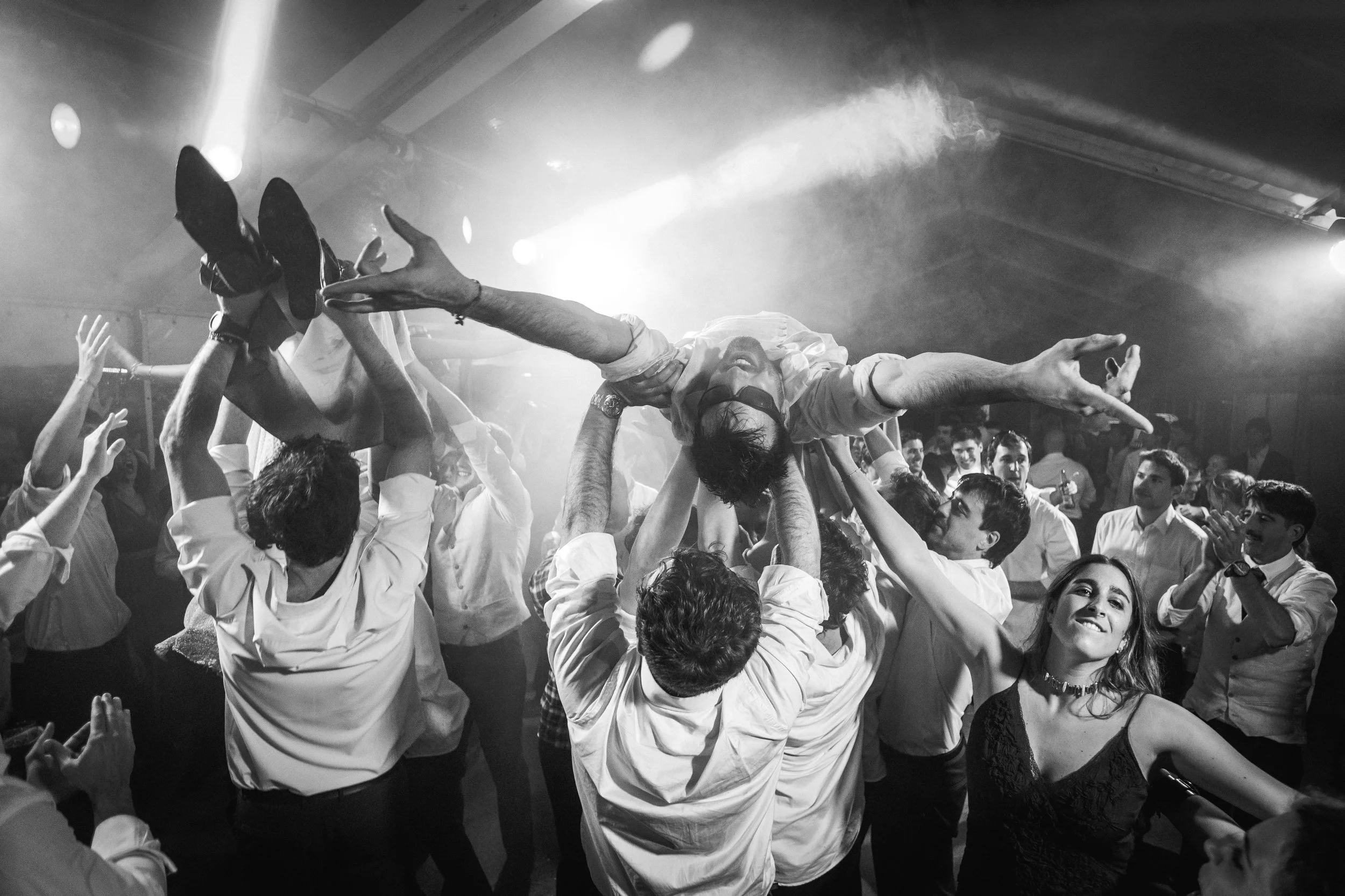 Diversas personas levantan a una persona en medio de un grupo en una celebración, con luces y humo en el fondo, en un ambiente festivo.