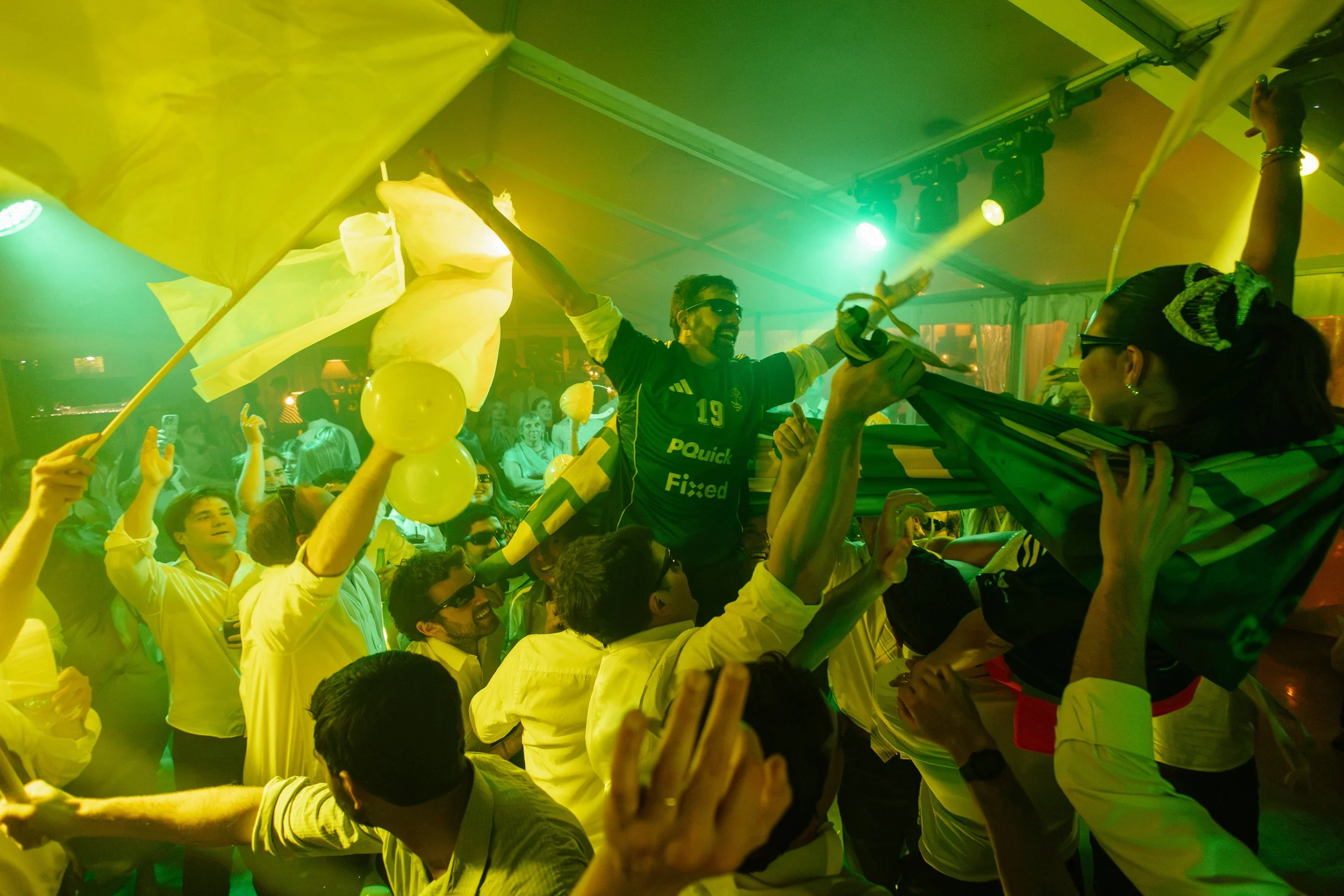 Grupo de personas celebrando en un evento con luces verdes y amarillas, levantando banderas y aire de alegría.