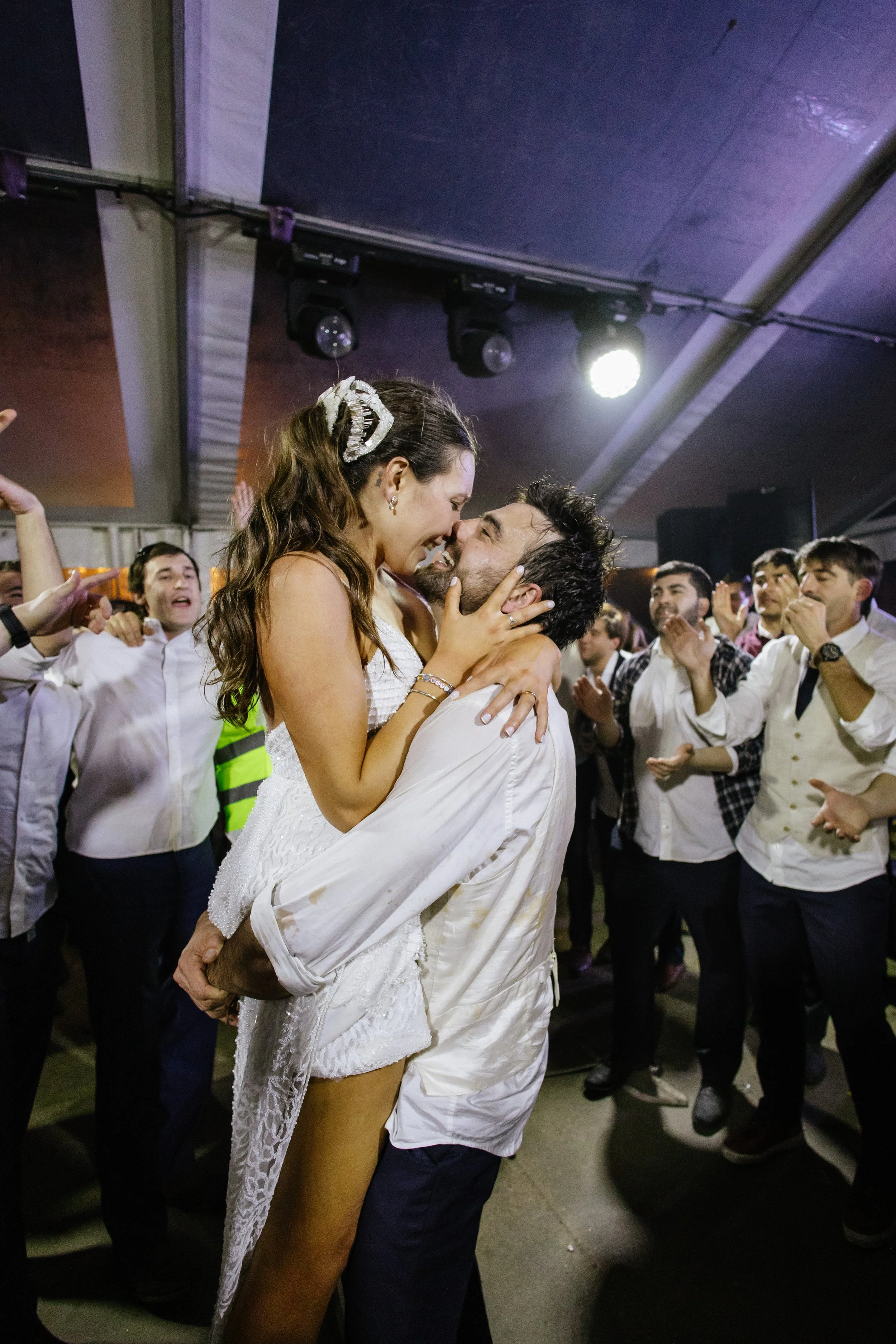 Pareja bailando en una celebración, rodeados de personas que los aplauden, en un lugar con iluminación de escenario.