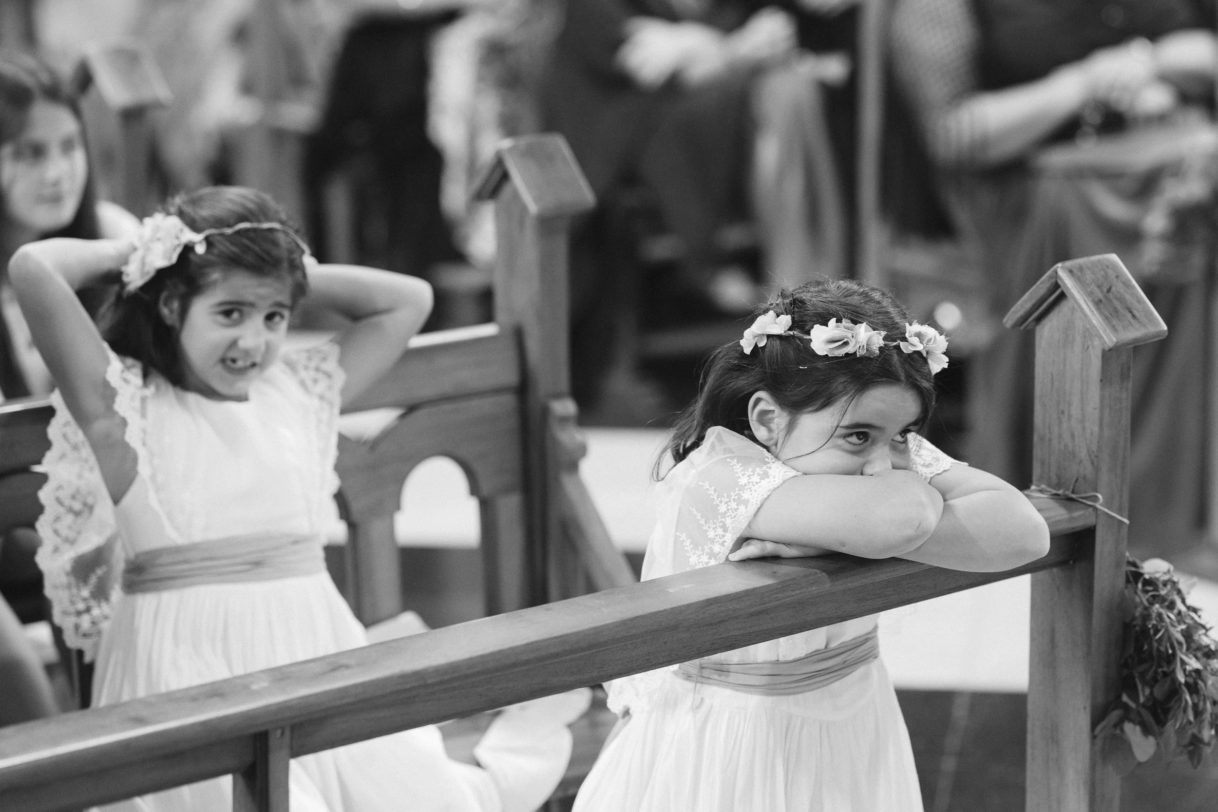 Niñas en una ceremonia religiosa, una de ellas con brazos cruzados sobre el mostrador y otra sonriendo, ambas vestidas con vestimenta blanca y diademas con flores.