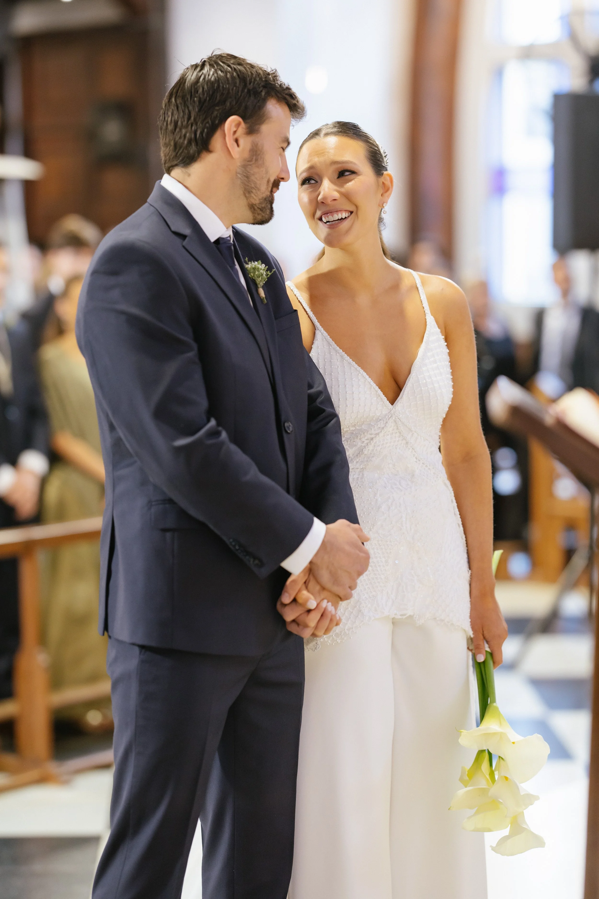 Una pareja en una boda, el hombre viste un traje oscuro y la mujer lleva un vestido blanco con tirantes y sostiene un ramo de flores blancas, están tomados de las manos y parecen felices en la ceremonia religiosa.
